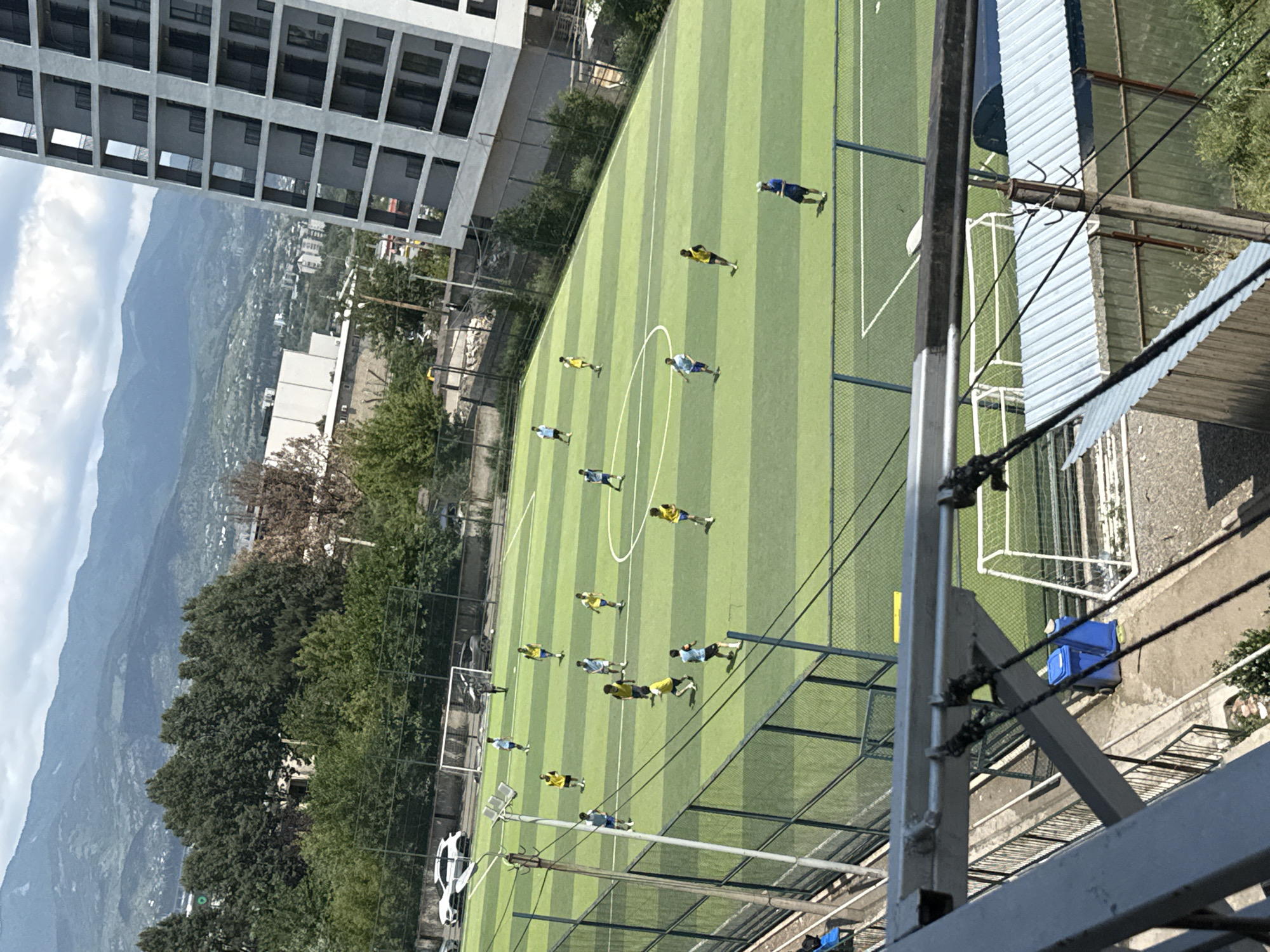 In Tbilisi, Georgia, a football match is underway on a bright, sunny day on an artificial turf field marked with green and white stripes. A group of young men, dressed in yellow and blue jerseys, are actively playing. One player in a blue jersey stands near the sidelines, observing the game. In the background, a tall, modern apartment building with numerous balconies rises against a backdrop of lush green trees and rolling mountains. The city's landscape is visible below the mountains, indicating an urban setting. The football field is enclosed by a dark fence, with a white goal structure visible on the left side of the field.
