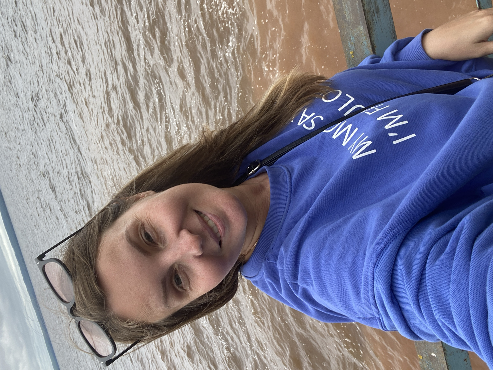 A woman with long brown hair is taking a selfie on a pier in Noviy, Russia. She is wearing a blue sweatshirt with white text that reads "MY MOSA I'M FABULOUS". Her sunglasses are perched on top of her head. The water behind her is murky brown and appears to be a river or bay. The sky is overcast and a railing runs along the edge of the pier.