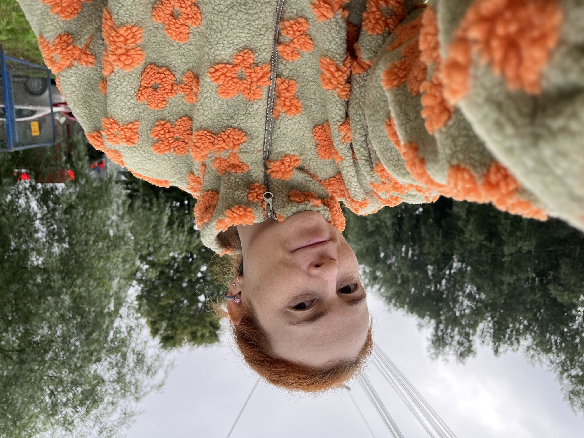 A woman with red hair takes a selfie outdoors. She is wearing a light green fleece jacket with orange floral embroidery. Her left arm is extended towards the camera, holding a device. She has a pink earbud in her right ear and a small earring in her left earlobe. In the background, there are trees and a cloudy sky, with visible power lines overhead. To the right, a blue fence encloses a parking area with a car partially visible. The setting appears to be an urban environment, likely Chaikovsky, Russia, given the presence of typical infrastructure like power lines and fencing.