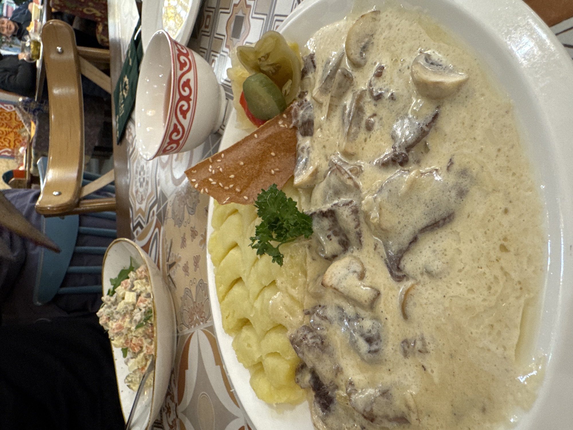 A close-up, slightly tilted view captures a hearty meal served on a patterned table, likely in a restaurant or cafe in Bishkek City, Kyrgyzstan. The central focus is a large white plate presenting a generous portion of a creamy mushroom and meat dish, resembling beef stroganoff, served alongside a textured mound of mashed potatoes. A sprig of fresh parsley garnishes the potatoes. Next to the main course, a triangular piece of flatbread or cracker, topped with sesame seeds, stands upright, accompanied by pickled vegetables including gherkins and what appears to be a pickled bell pepper or tomato.
To the left, a white bowl with distinctive red, swirling, traditional Central Asian patterns is visible. Further left and lower in the frame, a smaller, boat-shaped white bowl contains a side salad, possibly an Olivier salad, featuring diced vegetables and what looks like meat or cheese.
In the blurred background, parts of wooden chairs are visible, along with reflections on the table surface. A person's face, smiling and looking towards the camera, is partially visible in the upper left background, suggesting a social dining setting. Another person is partially visible, seated in the lower left. The table features intricate geometric and floral patterns, common in the region. There is no visible text in the image.