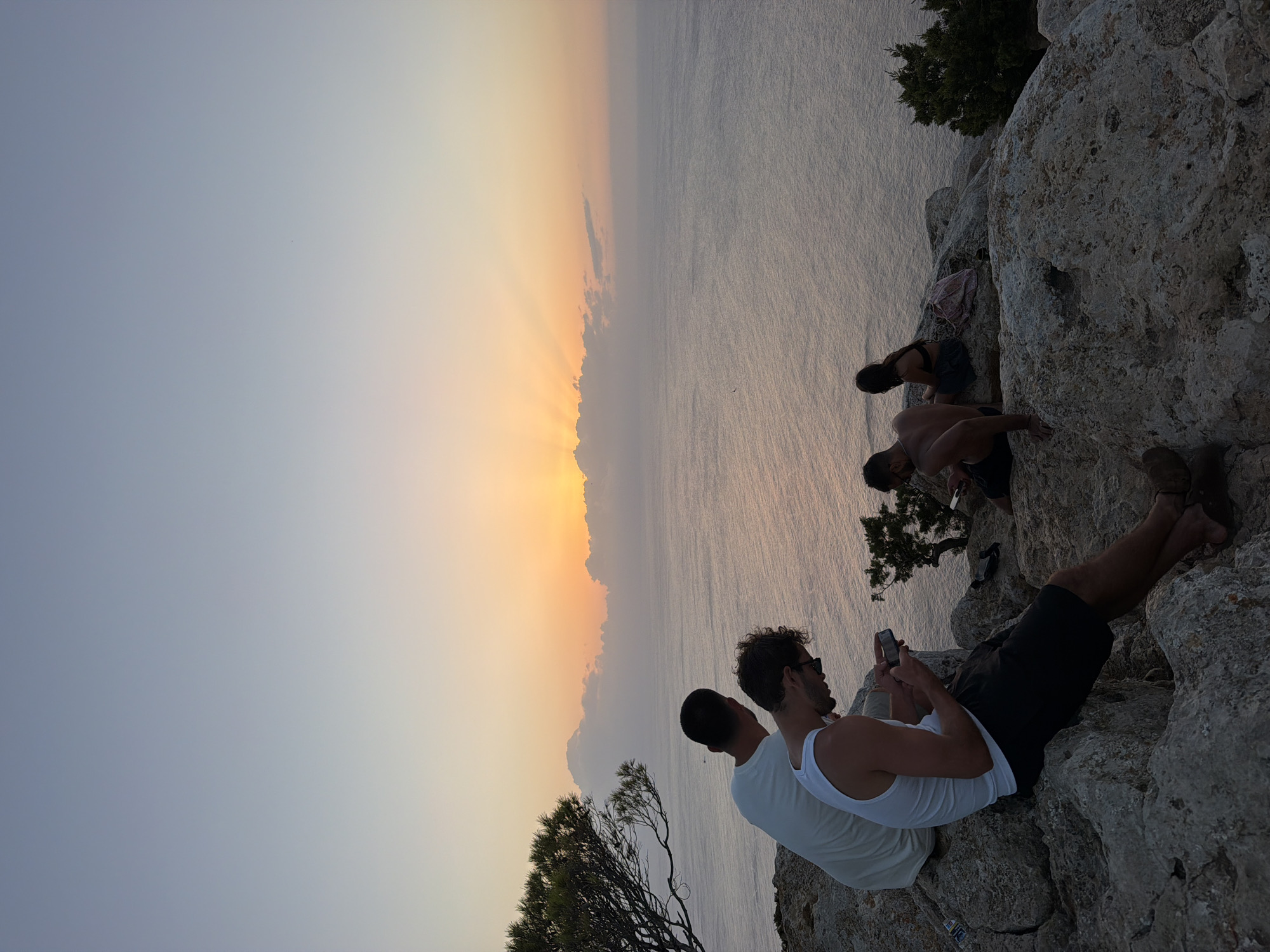 The image shows a group of people sitting on a rocky cliff overlooking the ocean at sunset. The sun is setting on the horizon, casting a warm glow over the water and sky. The people appear to be young adults, and they are dressed casually. Some of them are looking at their phones, while others are talking to each other. The scene is peaceful and serene.
The location is likely a viewpoint in Sant Antoni de Portmany, Spain, known for its beautiful sunsets over the Mediterranean Sea. The rocky terrain and the presence of pine trees are consistent with the landscape of the coastal areas around Sant Antoni de Portmany.