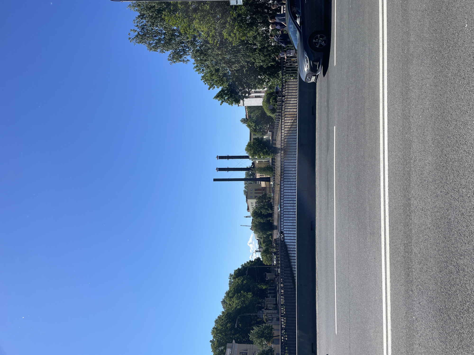 This image captures a sunny day scene in Saint Petersburg, Russia, likely along one of its many canals. In the foreground, a dark grey sedan is driving on a multi-lane asphalt road, indicated by white lane markers. The road is bordered by a raised concrete barrier. Beyond the barrier is a canal with blue water. On the far side of the canal, a black metal fence runs along the embankment, behind which are lush green trees. Further back, a row of buildings is visible, including a light-colored structure with windows and several industrial-looking chimneys. Several people are seen walking along the embankment. The sky above is a clear, bright blue.