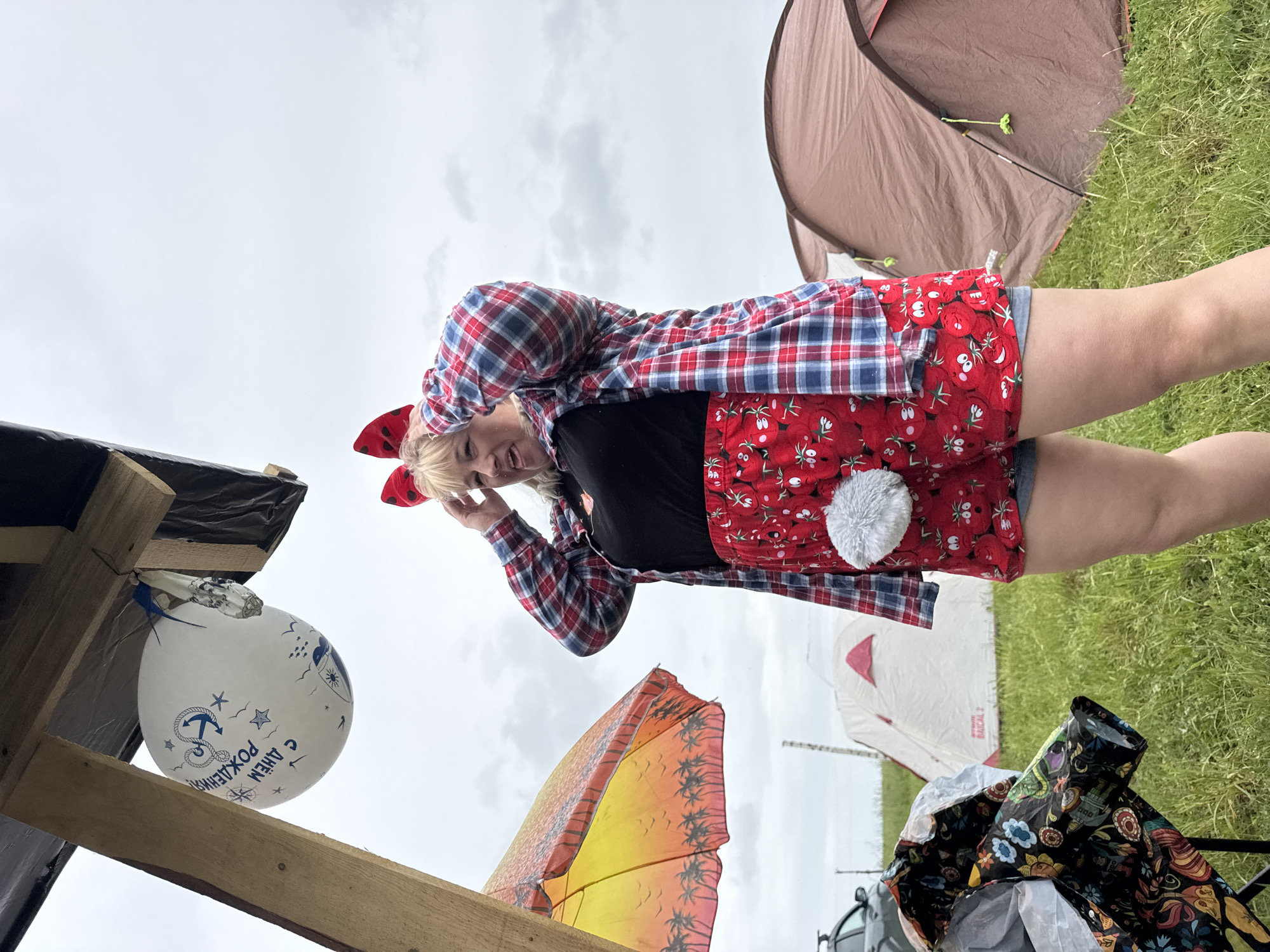 An outdoor scene captured from a low angle shows a woman smiling, possibly posing, against a cloudy sky. She is wearing a red, blue, and white plaid shirt, a black top, and red shorts or a short skirt adorned with a pattern of white faces on red fruit, featuring a white fluffy pom-pom attached. A red bow is visible in her blonde hair.
In the background, to her right, stands a brown, dome-shaped camping tent with green and black poles visible near its opening. To her left, there are wooden structural elements, possibly part of a makeshift shelter. A white balloon, partially obscured by these structures, hangs with blue marine-themed decorations including an anchor, stars, and waves. Visible text on the balloon reads "С ДНЁМ РОЖДЕНИЯ!", which translates from Russian to "Happy Birthday!". Below the wooden structure, a large orange and yellow umbrella with a palm tree silhouette pattern is visible.
Further in the background, a grassy field stretches out with another white tent partially visible, which has "NORDWAY" and "BALACLAVA 2" (or similar brand text) printed on it. The overall setting suggests a casual outdoor event or camping trip, possibly a birthday celebration, under an overcast sky in a natural environment. The woman's expression is one of contentment or amusement.