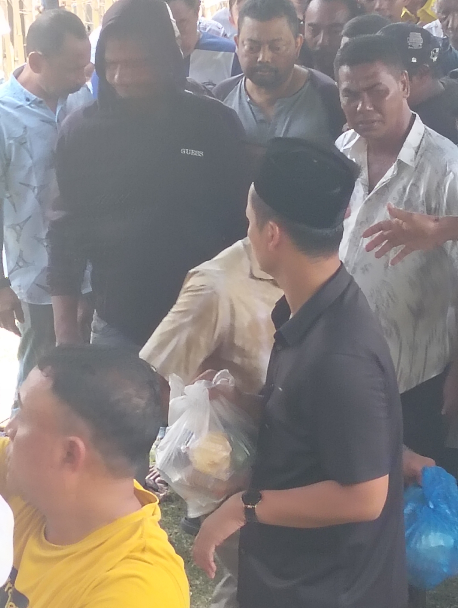 The image depicts a group of men in an outdoor setting, likely in Grong-Grong, Indonesia, based on the provided context. In the foreground, a man wearing a bright yellow t-shirt is visible from the chest up, his head turned away from the camera. To his right, a man in a black long-sleeved shirt and a black cap is seen from the shoulders up, holding a clear plastic bag filled with items. Behind him, another man in a light-colored, patterned short-sleeved shirt is partially visible, his hand reaching towards the man in the black shirt. Further back, a man in a black hooded sweatshirt with the "GUESS" logo is prominent. Other individuals are present in the background, forming a crowd. The ground appears to be grassy. The lighting suggests it is daytime.