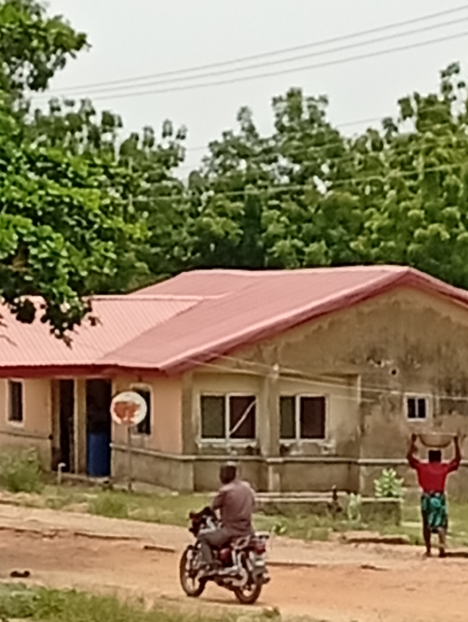 In Adankolo, Nigeria, a single-story building with a red roof occupies the scene. The building has multiple windows and a sign. A man rides a motorcycle on a dirt road in front of the building. A woman walks to the right of the building carrying something on her head. Trees line the background and power lines cross the sky.