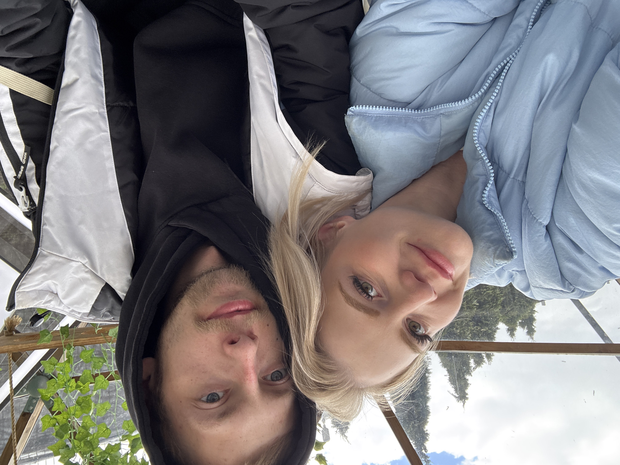This close-up portrait shows a young man and a young woman, likely a couple, posing for a selfie inside a transparent structure, possibly a greenhouse or conservatory. The image is rotated 90 degrees counter-clockwise.
The man, on the left, has light stubble, blue eyes, and wears a black hooded jacket with a white or light gray lining. He has a subtle, pleasant expression. On his jacket zipper pull, the text "WIND SPEED" is visible.
The woman, on the right, has blonde hair, green/hazel eyes with noticeable eyeliner and mascara, and wears a light blue puffer jacket. She displays a soft smile with pink-tinted lips. Their heads are close together, looking directly at the camera.
Above and around them, the transparent panels and wooden beams of a roof structure are visible. Green, leafy plants, possibly ivy, hang within the structure near the man. Through the glass, the background reveals a partly cloudy sky with patches of blue and distant green coniferous trees, suggesting an outdoor location.
The time of day appears to be daytime, under bright but partially overcast conditions. The setting, within a glass enclosure and with external trees, fits the context of a park or garden in Saint Petersburg, Russia. The overall mood is relaxed and intimate.