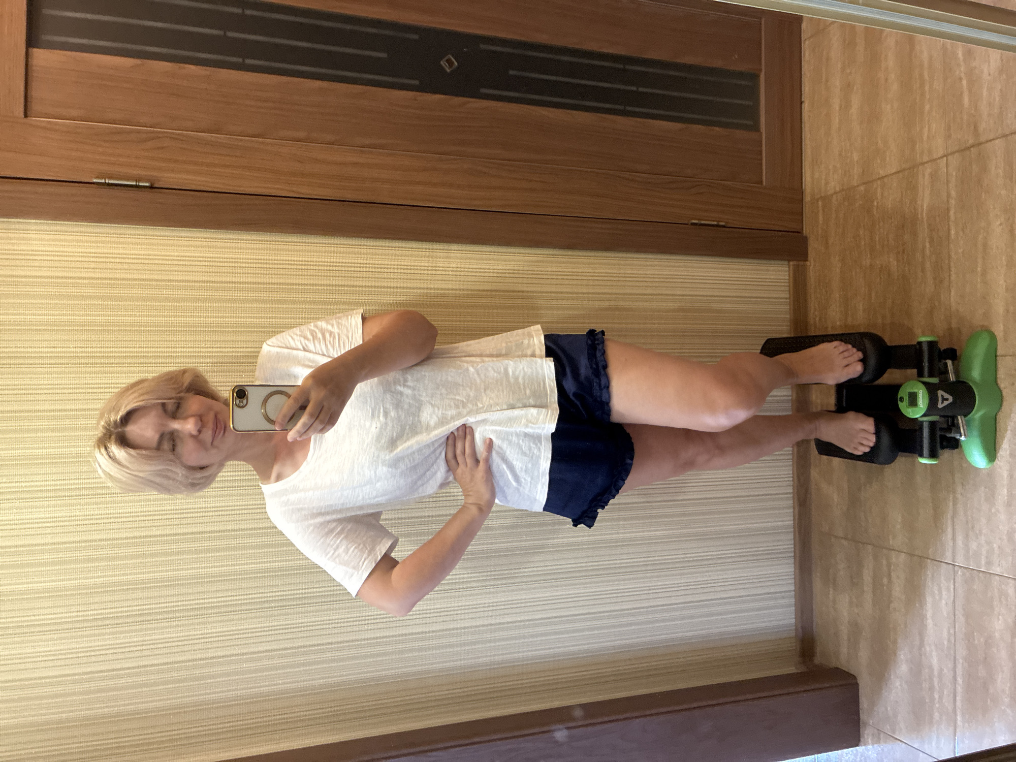 In Sochi, Russia, a woman with blonde hair stands indoors, taking a selfie in front of a wall covered in vertical striped wallpaper. She is wearing a white t-shirt and navy blue shorts with ruffled hems. Her right hand is on her hip. She is standing on a green and black exercise stepper. To her right is a wooden door with vertical dark panels. The floor is tiled in a light brown and cream pattern.