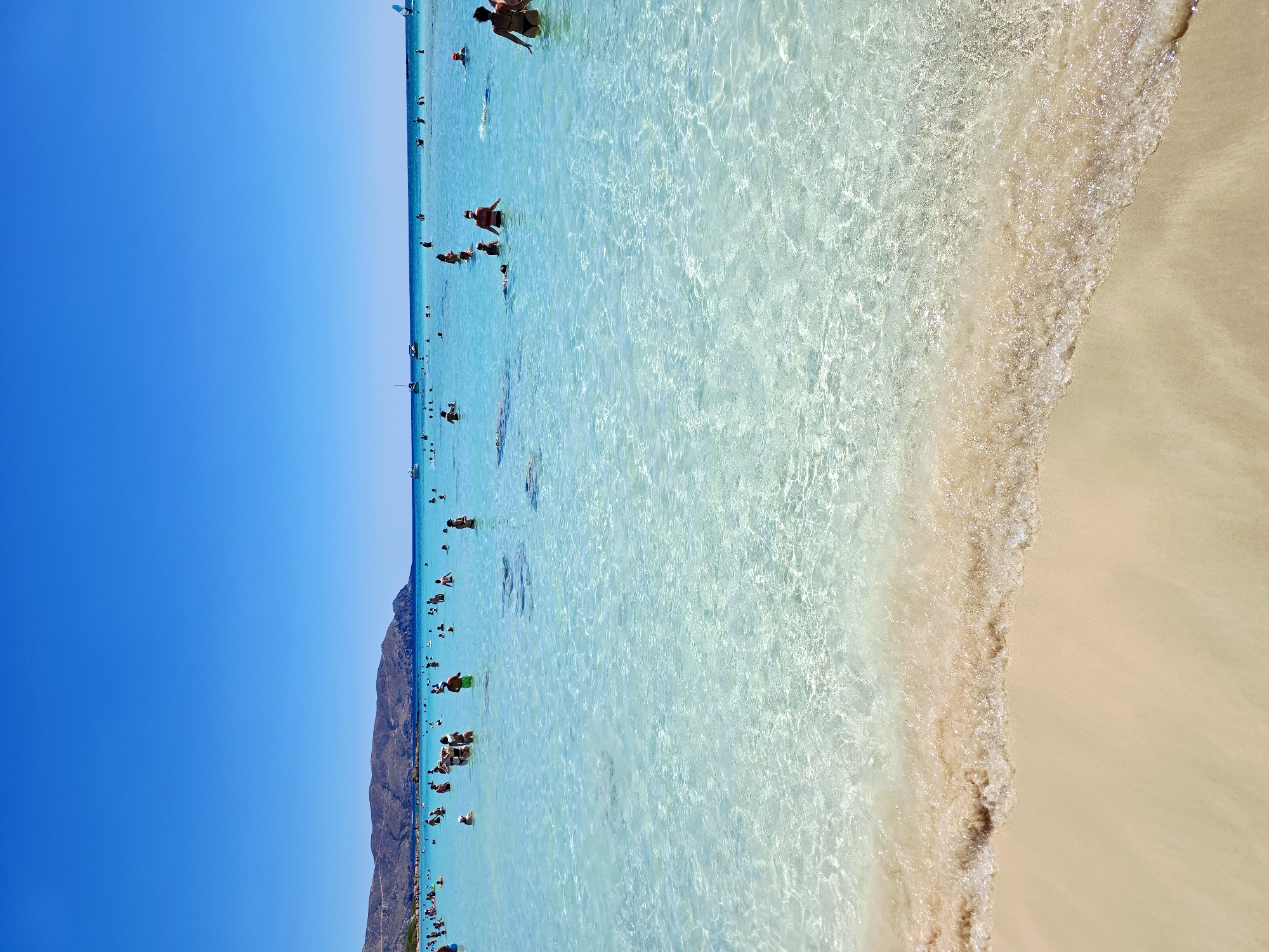 This image depicts a crowded beach scene in Greece, likely on a sunny day. The water is crystal clear and turquoise, with gentle waves lapping onto a sandy shore. Numerous people are visible in the water, swimming, wading, and enjoying the beach. Some are standing, while others are partially submerged. The background shows a hazy coastline with hills and mountains. The sky is a vibrant, cloudless blue. Several boats and a windsurfer are also present in the water. The overall impression is one of a popular tourist destination with active recreational use.