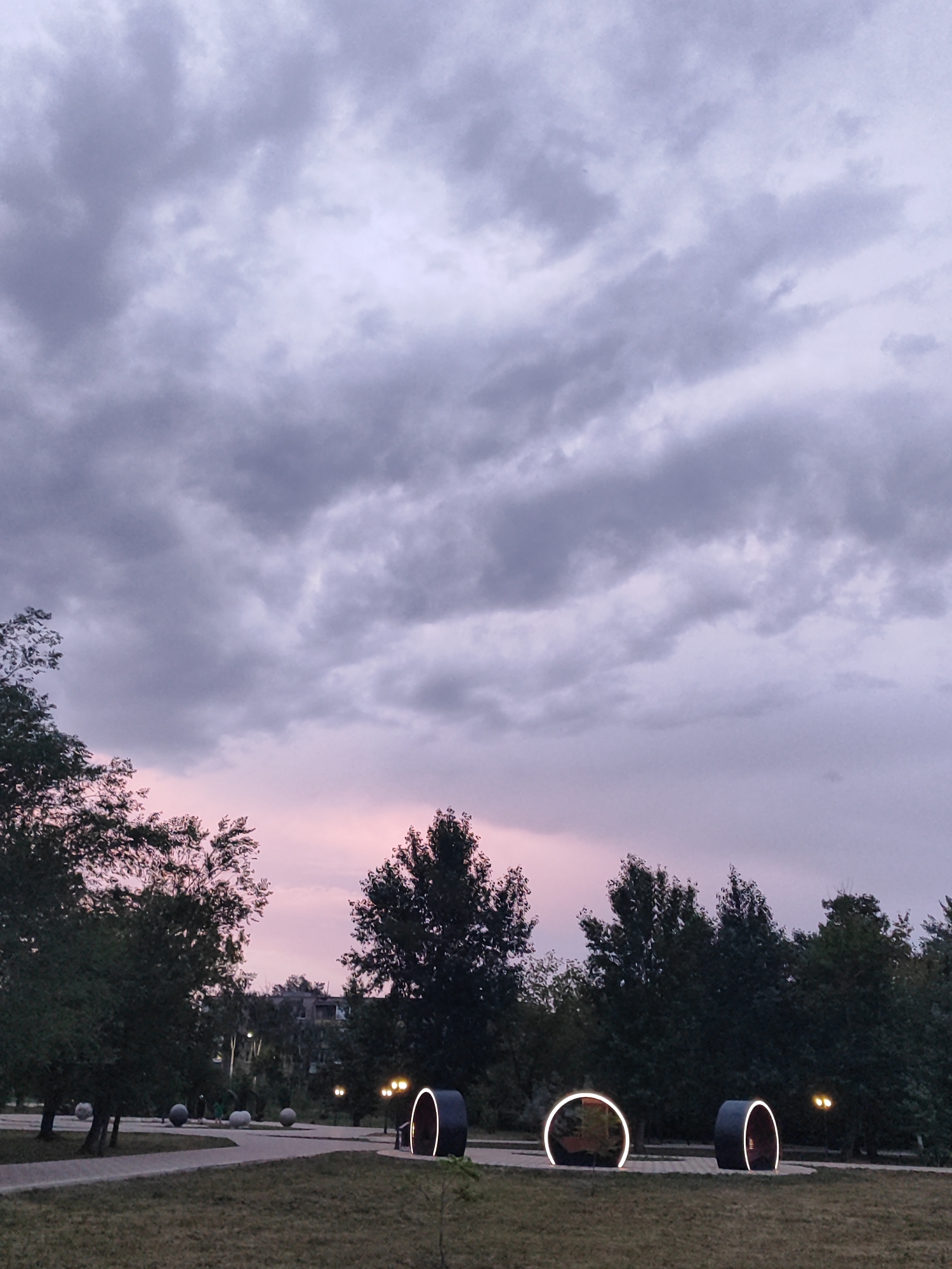 The image depicts a park scene at dusk in Slavgorod, Russia. The sky is filled with dramatic, dark gray clouds that are tinged with pink and purple hues from the setting sun. The lighting suggests it is twilight.
In the foreground, a grassy area transitions into a paved walkway that winds through the park. Scattered along the walkway are several large, spherical objects that appear to be decorative or artistic installations. Further into the park, there are three distinct, modern, circular sculptures, each illuminated with white light, creating a striking contrast against the darkening landscape. Two of these sculptures are large, open rings, while a third, to the right, is a more solid, tunnel-like structure.
Behind the sculptures and walkways, a dense line of dark green trees forms the background. Beyond the trees, the silhouette of buildings can be seen, suggesting the park is located in or near a populated area. Several streetlights are visible, casting a warm, yellow glow. The overall mood is serene and slightly moody, with the dramatic sky and the illuminated sculptures creating a sense of quiet wonder. There are no people visible in the image. The weather appears overcast, typical of evening skies.