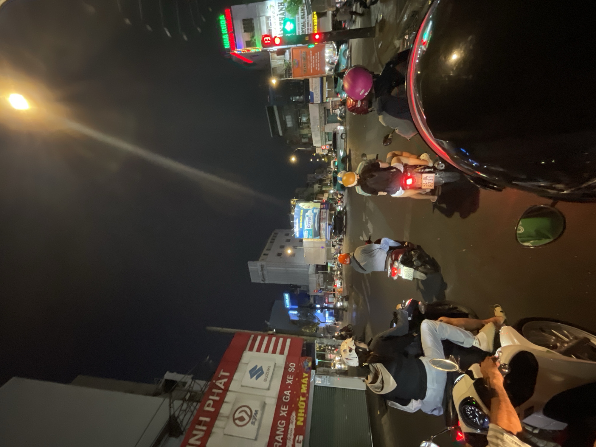 This image, taken at night in the city of Thủ Đức, Vietnam, depicts a bustling street scene with numerous scooters and motorcycles navigating the roadways. The foreground is dominated by a motorcycle with a dark helmet. People are riding the scooters, some wearing helmets. The street is lined with various businesses and advertisements, including signs for "SYM" and "SUZUKI," along with the phrase "NHỚT MÁY". In the distance, there is a lit-up sign for "KEM ĐÁNH RĂNG Doreen" (Doreen Toothpaste). There are also signs for "EXIMBANK" and "NHA KHOA". Traffic lights and street lamps illuminate the area, casting shadows and highlighting the activity on the road.