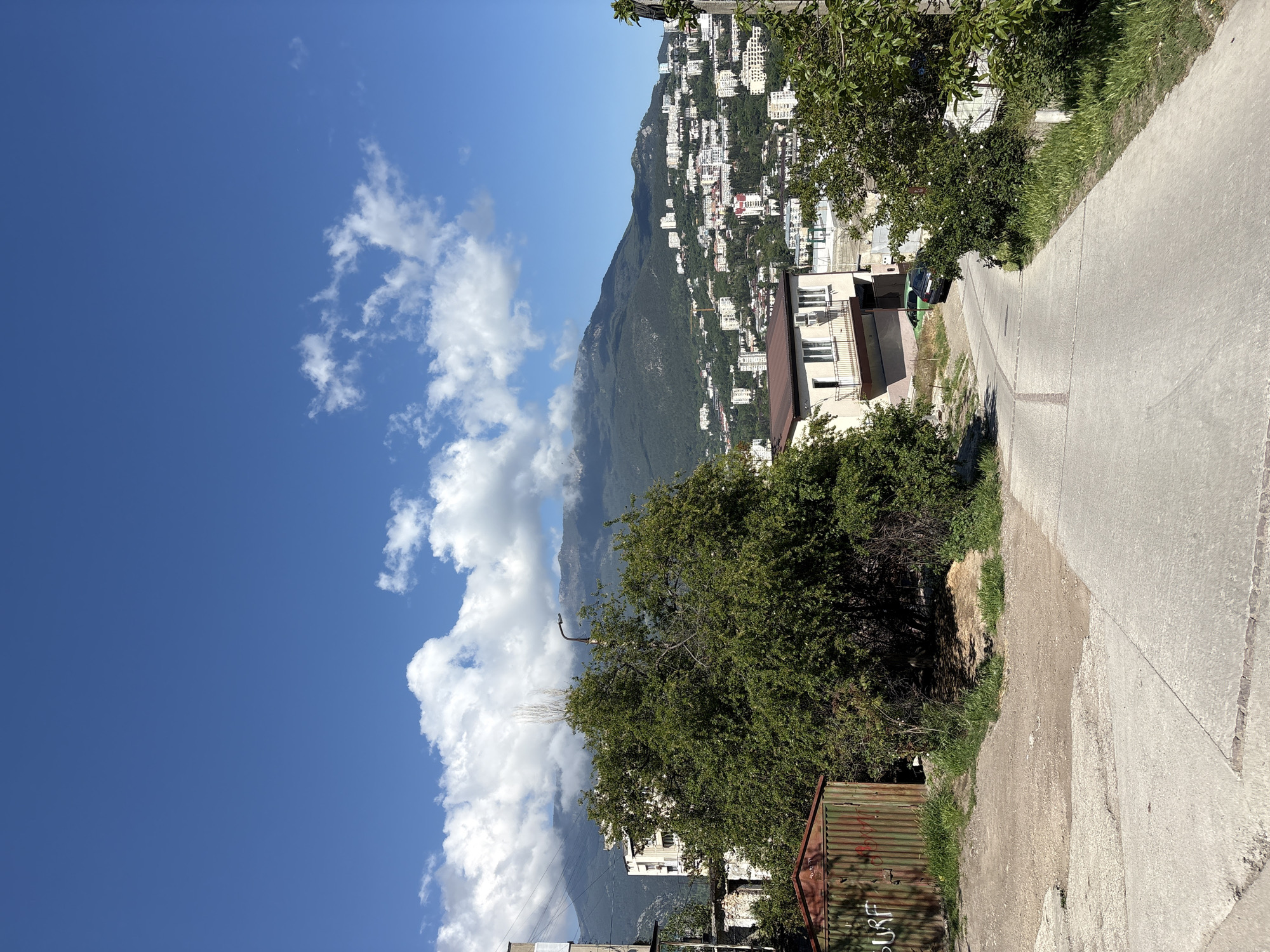 A photograph depicts a hillside in город Ялта, Ukraine, densely populated with white buildings and vegetation. A concrete road runs along the right side of the frame. A tree and a corrugated metal structure occupy the lower right corner. The left side of the frame is filled with a bright blue sky and white clouds, while a mountain extends between the sky and the city.