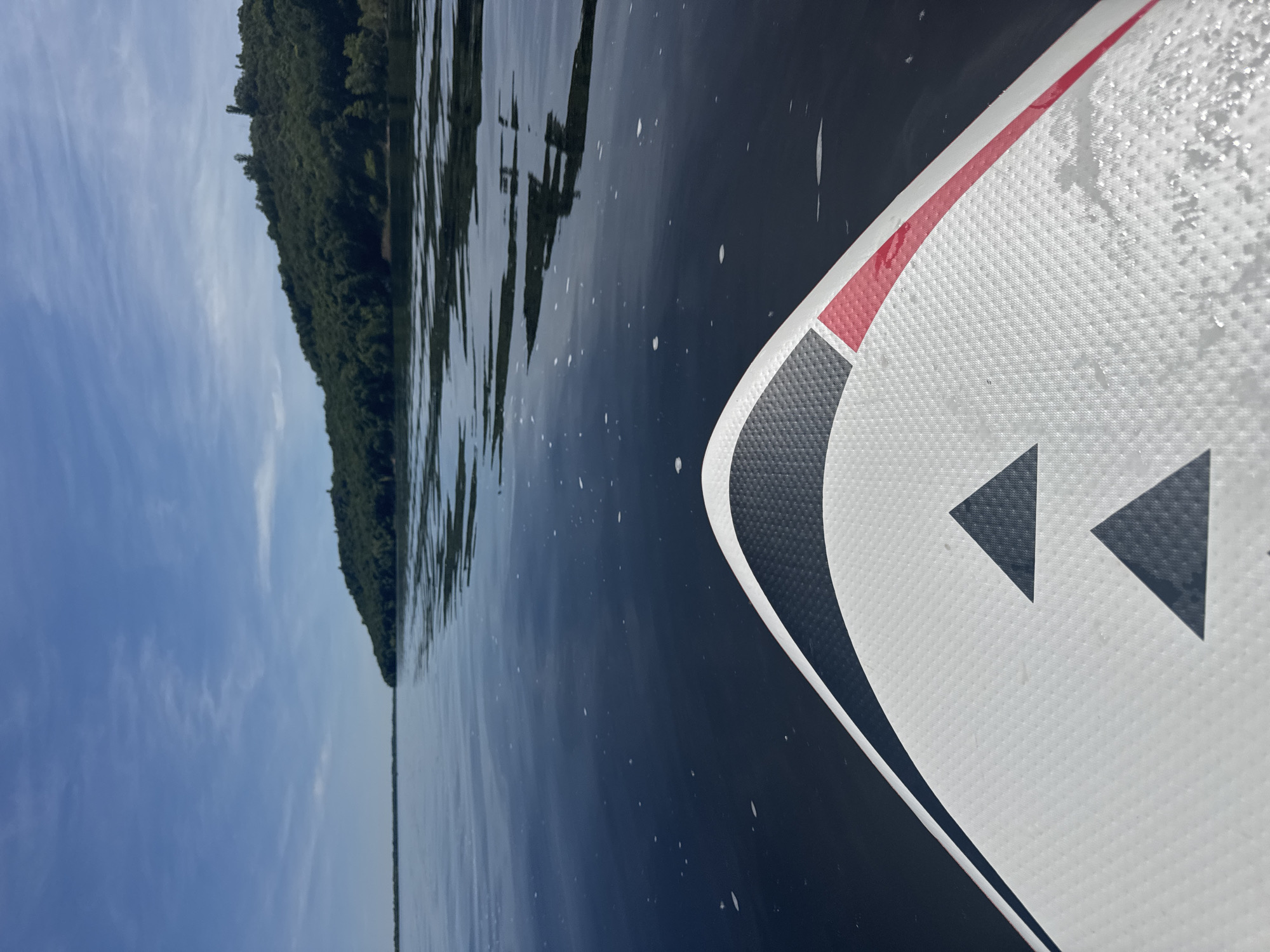 An eye-level shot captures a serene view from the bow of a stand-up paddleboard or kayak on a calm body of water. The image is oriented vertically, showing the watercraft in the lower right foreground, pointing towards a distant forested shoreline under a clear sky.
The watercraft's textured, non-slip deck is primarily white, adorned with a design featuring a prominent red stripe, black and white curved sections, and two dark triangular shapes, possibly decorative or functional markers. Small water droplets are visible on the surface of the board, suggesting recent movement through the water.
The water is dark and reflective, mirroring the distant trees and showing subtle ripples. Specks of foam or tiny bubbles are scattered across its surface, especially near the board. On the left side of the frame, the dark green canopy of a dense forest forms a low, continuous line along the horizon, indicating a distant shore. Above the forest, a clear blue sky extends upwards, with thin, wispy white clouds providing texture.
The scene depicts a peaceful outdoor activity on a bright, sunny day, likely during morning or afternoon hours given the natural light. The setting is identified as Усть-Курдюм, Russia, suggesting this tranquil experience is taking place on the Volga River or a connected waterway. No discernible text is visible in the image.