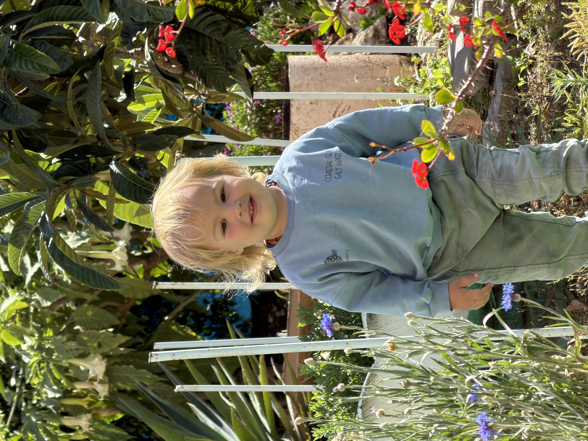 A young child with blond hair is standing in a garden, wearing a blue long-sleeved shirt and green pants. Red and blue flowers are visible in the foreground and background. The child is smiling and looking at the camera. A white metal fence is present. The location is possibly Kiryat Bialik, Israel.