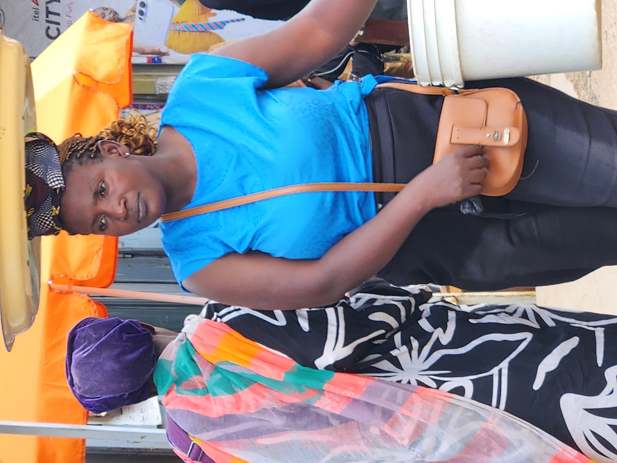 In an outdoor urban environment, two women are depicted. The woman on the right, wearing a blue top and black pants, is facing forward and carrying a small brown purse. She is standing next to a white bucket. The woman on the left, wearing a purple headscarf and a colorful patterned garment, is facing away from the camera. An orange awning is visible above them. The background includes a building and some signage.