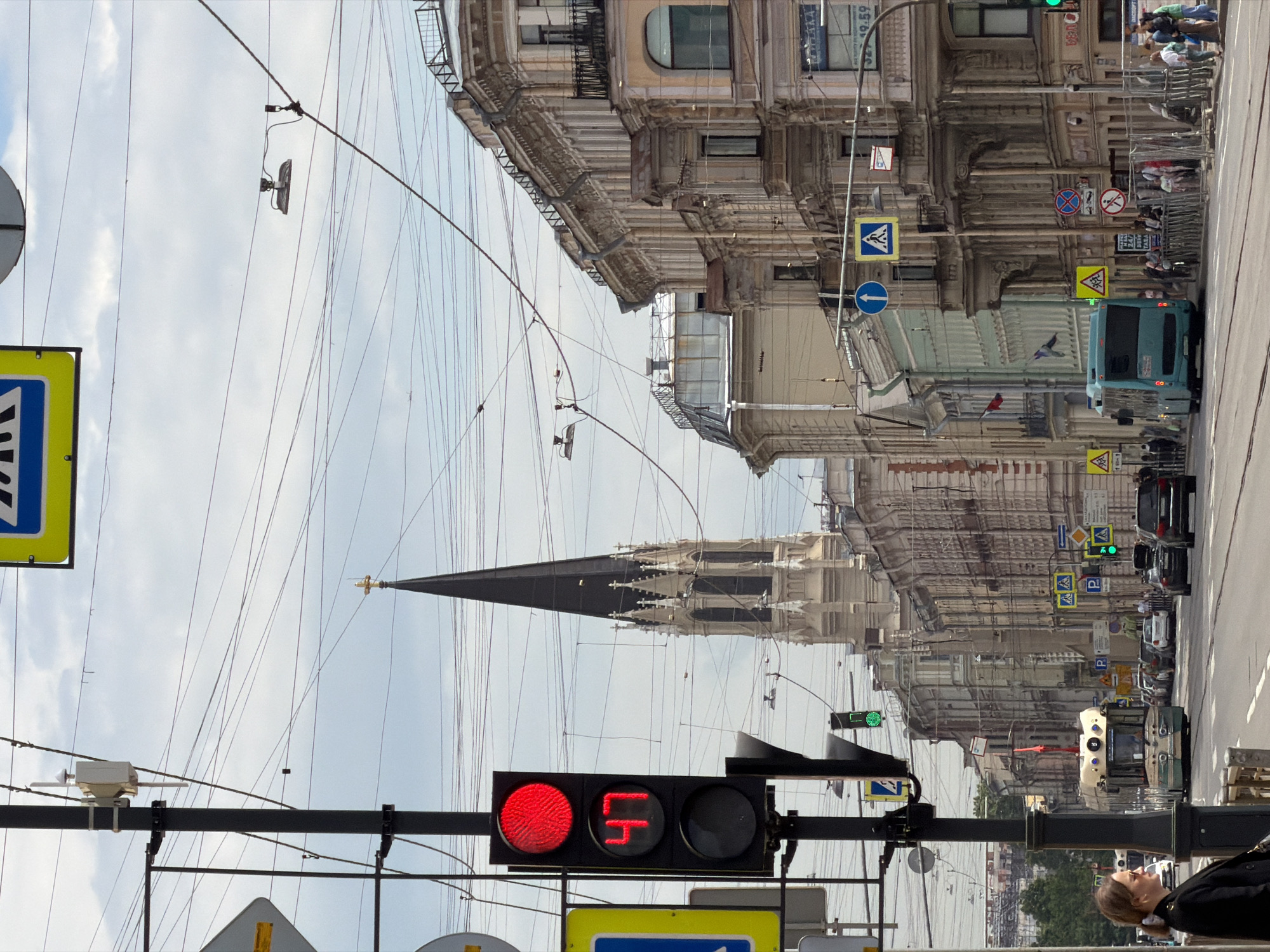 This image, rotated approximately 90 degrees counter-clockwise, captures a daytime street scene in Saint Petersburg, Russia. The view looks down a wide street flanked by ornate, multi-story classical buildings featuring balconies and intricate architectural details. A dense network of overhead wires, likely for trams or trolleybuses, crisscrosses the cloudy sky.
In the mid-ground, a tall, dark church spire with white architectural accents rises above the cityscape, a prominent landmark. In the immediate foreground, a traffic light shows a solid red circle and a red countdown display reading "Ч" (the Cyrillic letter for 'Ch' which looks like a '4'). Another traffic light further down the street is green. A person with light hair and a dark jacket is visible on the right side of the foreground.
The street is busy with vehicles, including a blue/green bus, several cars, and what appears to be a light-colored tram or trolleybus in the distance. Numerous road signs are present, including pedestrian crossings (both blue square and yellow diamond variants), a blue circular arrow pointing right, no parking/stopping signs, and yellow diamond signs warning of children. Some illegible text and numbers are visible on buildings, along with "БАР" (BAR in Cyrillic) and partially "327 19.59". The overall impression is a bustling urban environment under a bright, cloudy sky, reflecting typical daily activity in a historic city.