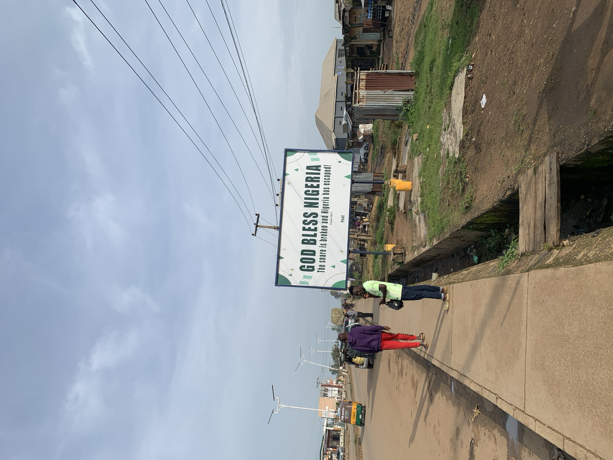 The image shows a street scene in Rafin Pa, Nigeria. A large billboard in the foreground reads "GOD BLESS NIGERIA. The snare is broken and Nigeria has escaped! Psalm 124:7 PoSe". Several people are visible, including a man in a purple hoodie and red pants walking away from the camera, and another man in a green shirt standing on the sidewalk. A three-wheeled vehicle, commonly known as an 'okada' or 'keke', is driving down the street. The environment appears to be somewhat underdeveloped, with some corrugated metal structures and uneven ground visible. The sky is overcast with some clouds. The street is paved, and there is a sidewalk present. Power lines are visible overhead. The overall impression is one of a typical street in a Nigerian town or city, with a focus on the billboard's message of hope or celebration.