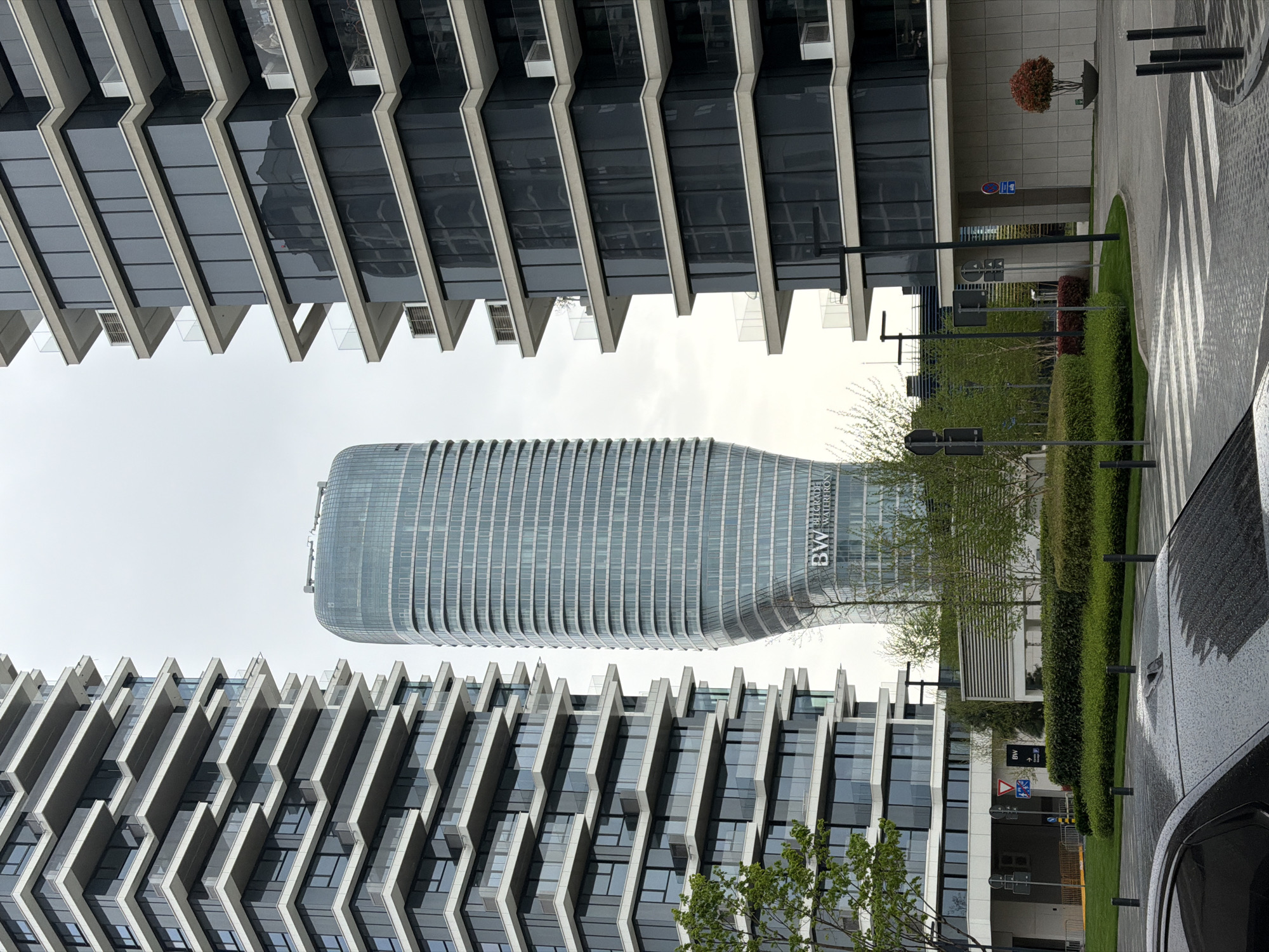 The image shows a view in Belgrade, Serbia, specifically in the Belgrade Waterfront area. The architecture features modern buildings with a distinct design of protruding balconies or sunshades. A uniquely shaped tower, labeled "BW Belgrade Waterfront," stands in the background. The foreground includes a street with a crosswalk, landscaping with bushes and trees, and the side of a black car. The overall scene appears to be taken on a slightly overcast day.