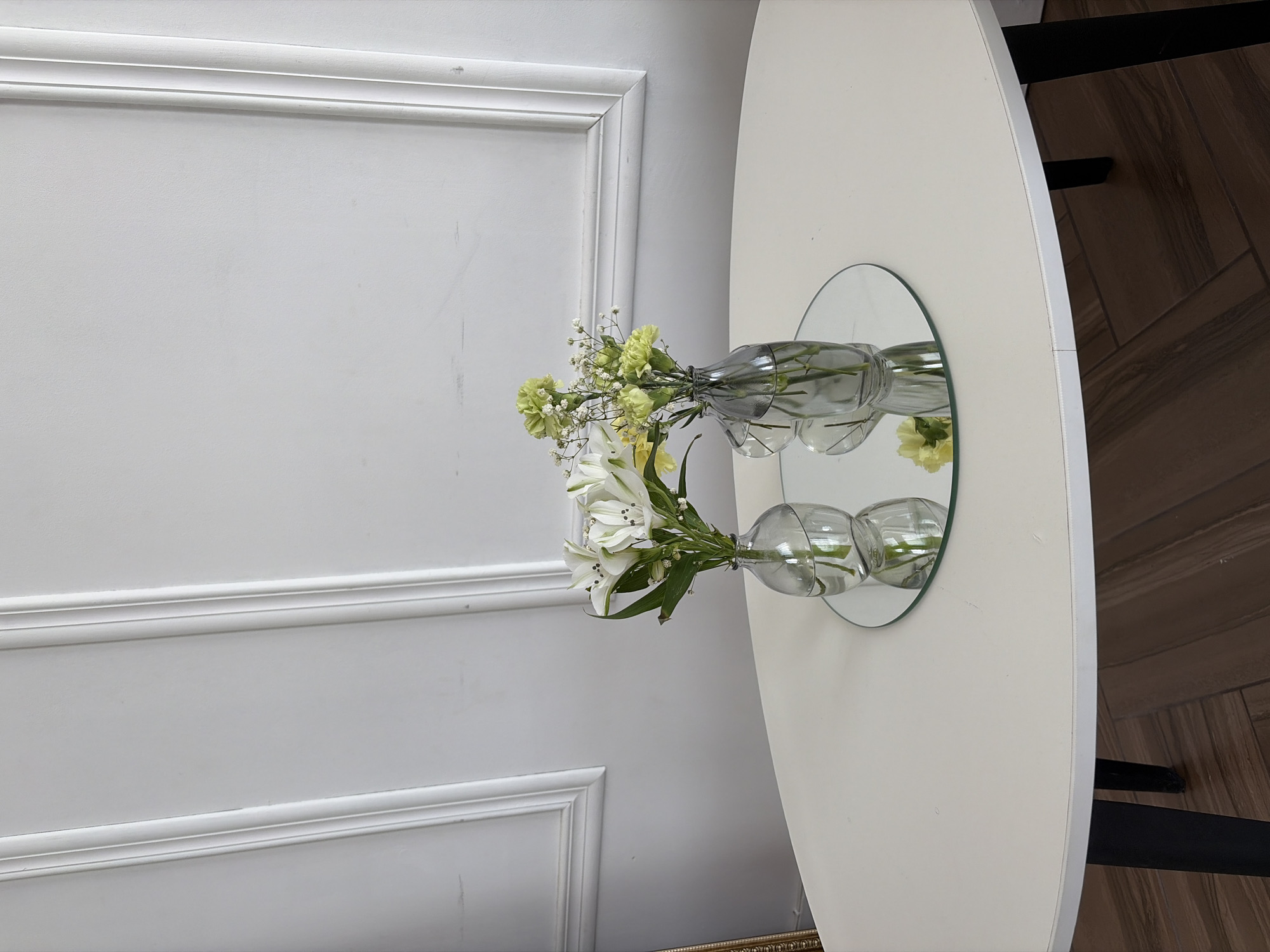 A vase of flowers sits atop a white, oval-shaped table with a mirrored surface, reflecting the floral arrangement and the table itself. The table is positioned against a light gray wall with framed paneling. The setting suggests an interior space. The location is Chaikovsky, Russia. The scene includes a vase, flowers, table, mirror, and a wall.