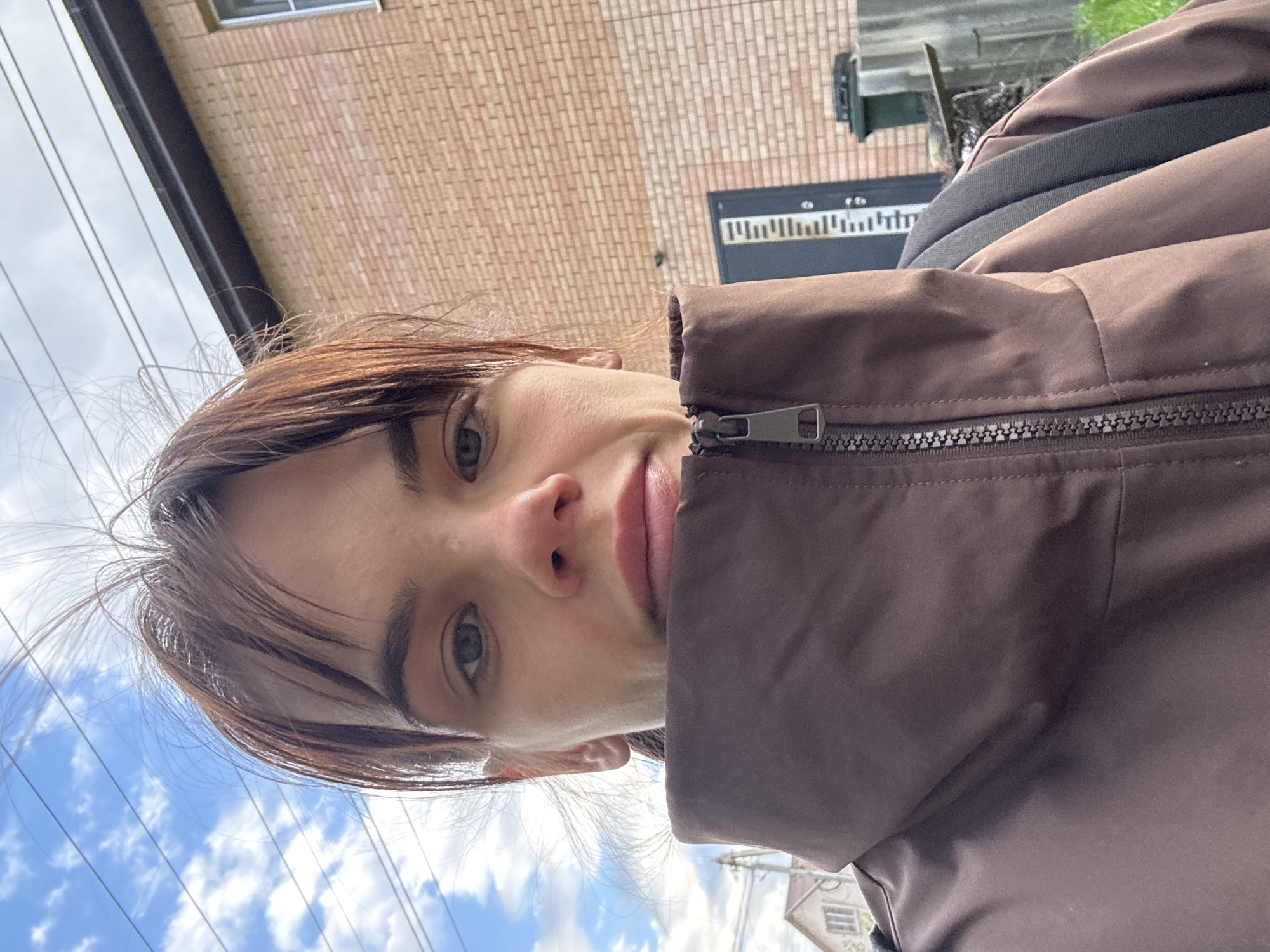 This is a vertically oriented, close-up selfie of a young person, rotated 90 degrees counter-clockwise. The individual has shoulder-length, light brown hair, which appears slightly windswept, and striking green or hazel eyes. They are wearing a brown, zippered puffer jacket, with the collar pulled up and partially obscuring the lower part of their face. Their expression is neutral, looking directly at the camera.
The setting appears to be outdoors during daytime, indicated by the bright, cloudy blue sky visible in the background. Electrical wires crisscross the sky towards the left. To the person's right (which is top right in the rotated image), a brick building with a window is visible. Further back, there's a dark, rectangular object or sign with faint white markings resembling a series of vertical bars or an equalizer display. Another building or structure is partially visible in the distance towards the bottom left of the frame. The weather seems fair, with soft, diffused sunlight.
The activity is clearly a self-portrait, captured from a slightly upward angle. The image provides context of Rostov-on-Don, Russia. No readable text is clearly discernible, only the pattern on the dark sign.
