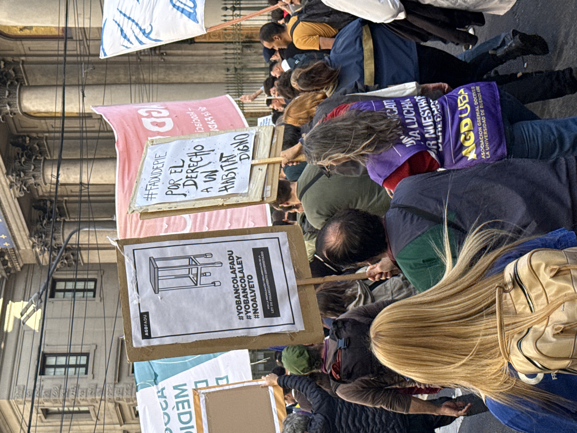 A medium shot of a crowd of protesters in Buenos Aires, Argentina, at an outdoor rally. The protesters are holding signs with text and drawings, including a sign with the text "#YOBANCOLAFADU #YOBANCOLALEY #NOALVETO" and a drawing of an upside-down chair; a sign with the text "#FADUDEPIF POR EL DERECHO A UN HABITAT DIGNO"; and a sign with the text "AGDUBA ASOCIACION GREMIAL DOCENTE DE LA UNIVERSIDAD DE BUENOS AIRES." Other people in the crowd wear shirts or carry banners with the text "UNICATO." The building in the background is a classic, ornate stone structure with windows, columns, and sculpted details. Power lines are visible overhead.
