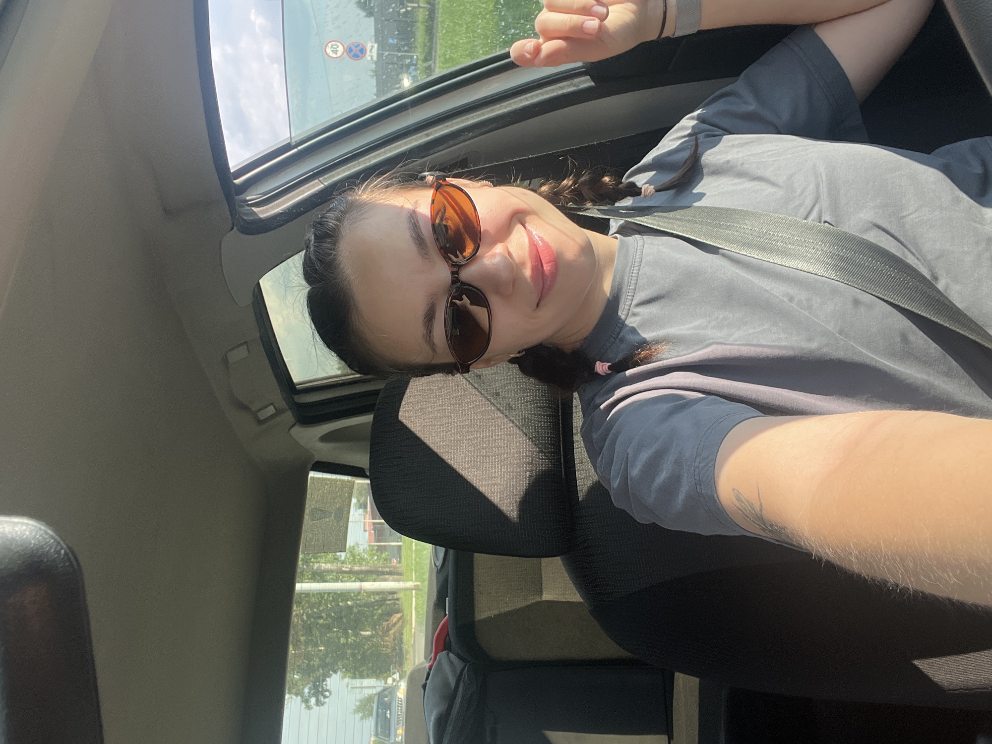 A young woman with brown hair in braids, wearing sunglasses and a gray t-shirt, is taking a selfie in the driver's seat of a car. She is smiling and her right hand is visible, raised slightly. The seatbelt is fastened across her chest. Sunlight is casting shadows inside the car. Outside the car, through the window, a street scene is visible with trees, buildings, a road, and a traffic sign. The location is identified as Chaikovsky, Russia.