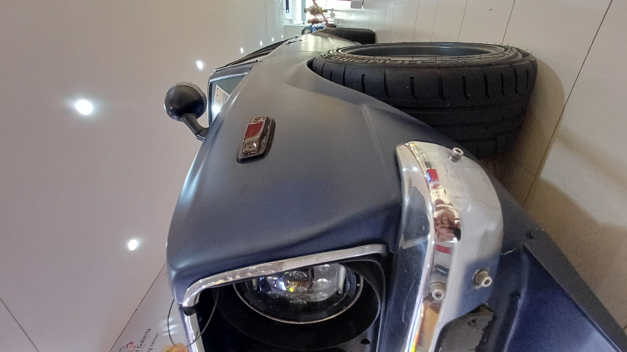 A cropped close-up shot of the front of a matte blue classic car, displaying a headlight with a chrome bezel, a black side mirror, a small red rectangular side marker light, and a large, stacked racing tire. The car is located inside of a gallery in the city of Kuta, Indonesia. The lighting is bright. A reflection of people can be seen in the chrome on the front of the car. The background is a light tan tiled floor and white walls with mounted lights.