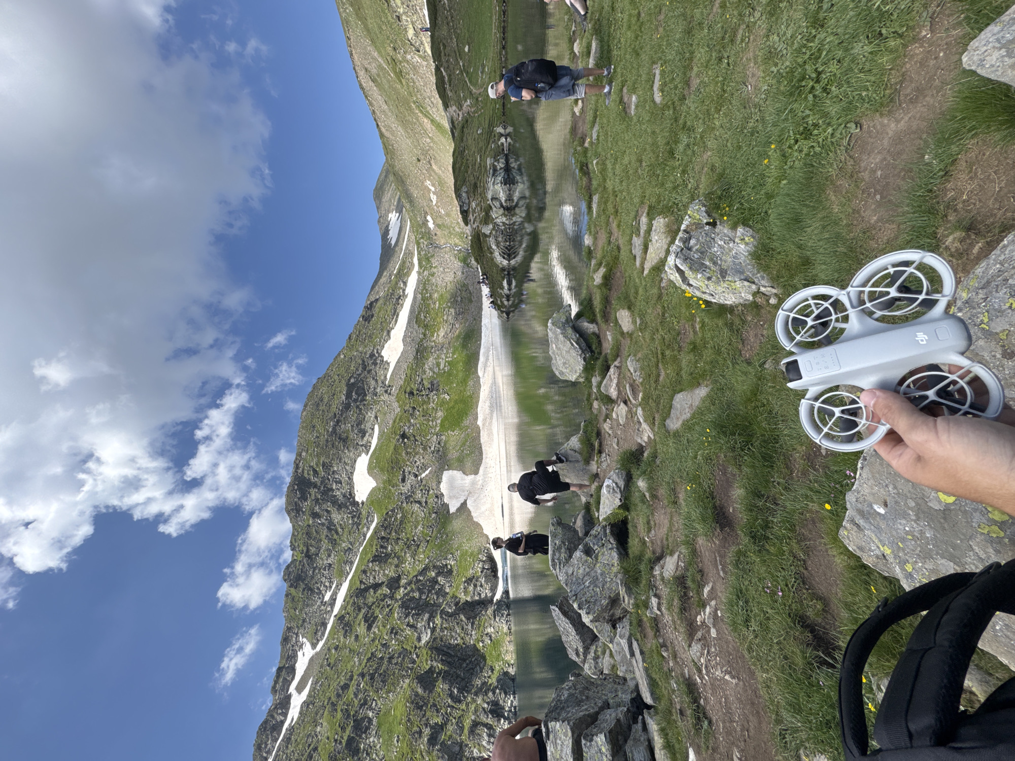 A drone is held in the foreground by a person's hand, positioned in front of a backpack. The drone is white and appears to be a DJI brand. In the background, a picturesque mountain lake is visible, with steep, rocky slopes covered in patches of snow and green vegetation. Several people are scattered around the lake, some near the water's edge and others on the grassy slopes. The sky is blue with scattered white clouds. The scene is consistent with the Seven Rila Lakes area in the Rila Mountains, near Panichishte, Bulgaria.