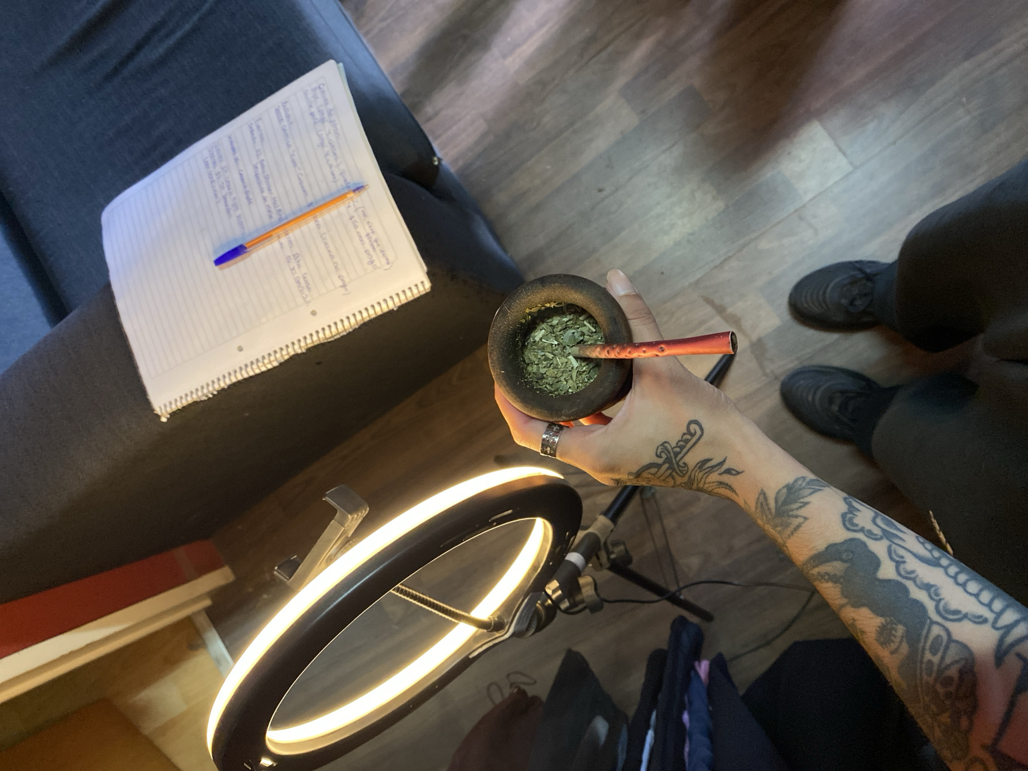The scene is an indoor setting, likely a home, based on the presence of a grey sofa, wood-patterned flooring, and a ring light. The location is stated as Llavallol, Argentina. A person's arm, heavily tattooed, is holding a traditional South American gourd (mate) filled with yerba mate. A bombilla (metal straw) is inserted into the gourd. The person is wearing a silver ring. To the left of the gourd is a notebook filled with handwritten notes and a pen. A ring light is positioned in the foreground. Black shoes are visible in the background.