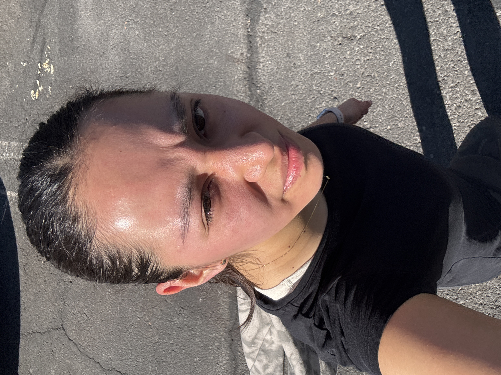 An angled, high-angle selfie captures a person outdoors against an asphalt background in bright sunlight. The subject is a person, likely female, with dark hair pulled back and appearing slicked or wet, possibly from sweat. Her face is turned upwards towards the camera, with a neutral to slightly soft expression. Her forehead, cheeks, and nose show a noticeable sheen, indicating perspiration. Her lips are slightly parted, revealing a hint of natural pink color.
She is wearing a black short-sleeved t-shirt, with a thin gold chain visible around her neck and a white layer (possibly an undershirt) peeking out at the collar. On her left wrist, a light-colored wristband or smartwatch is visible.
The background is a dark gray, textured paved surface, likely asphalt or concrete, with some lighter specks and cracks. Prominent, elongated, dark shadows are cast diagonally across the ground, suggesting strong, direct sunlight, indicative of either morning or late afternoon. No specific objects other than the ground and shadows are discernible in the background. No text is visible in the image.
The overall scene conveys an outdoor setting, potentially after or during some physical activity given the apparent sweat, on a sunny day in Puebla, Mexico.