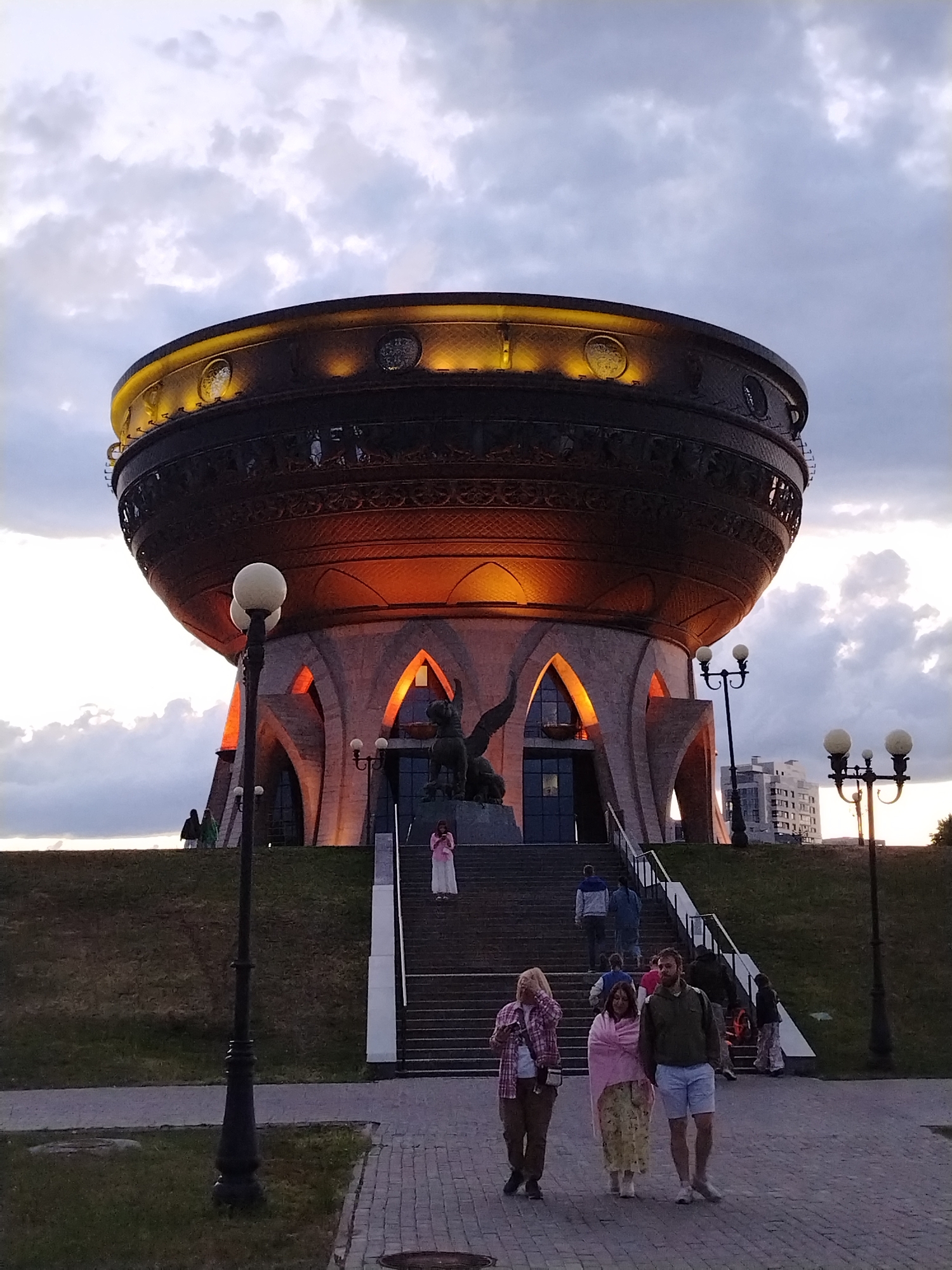 The image captures an evening scene featuring the distinctive Kazan Family Center (ZAGS) in Kazan, Russia, often referred to as "the Bowl" or "Chasha." The central subject is the colossal, dark bronze-colored building, shaped like a giant kazan (a cooking pot), which dominates the upper half of the frame. It is ornately detailed with intricate patterns and lit from within its upper rim with warm yellow lights. Below the bowl, the structure is supported by four large, arched pillars, which are illuminated with a soft orange glow, creating a dramatic effect against the twilight sky. At the base of the building, on a large set of stairs, a statue of a mythical winged creature (likely a Zilant, a symbol of Kazan) is visible near the main entrance.
In the foreground, a paved pathway leads towards the building. Three people are walking away from the camera on this path: a woman with light-colored hair and a plaid shirt, a woman in a long patterned dress and a pink shawl, and a man in a green jacket and light-colored shorts. Further back, several other individuals are seen ascending the wide stone staircase that leads up to the building's entrance. On the right side of the stairs, another person is standing, possibly looking at a phone. More people are visible on the grassy incline to the left.
The time of day appears to be dusk or early evening, indicated by the soft, diffused light from the cloudy sky and the artificial illumination of the building. The clouds are a mixture of grays and whites, suggesting an overcast but calm sky. Two prominent street lamps with spherical lights stand on either side of the pathway in the foreground. A modern building is partially visible in the background on the right. There is no clearly legible text visible in the image. The overall atmosphere is serene, with visitors casually exploring the architectural landmark.