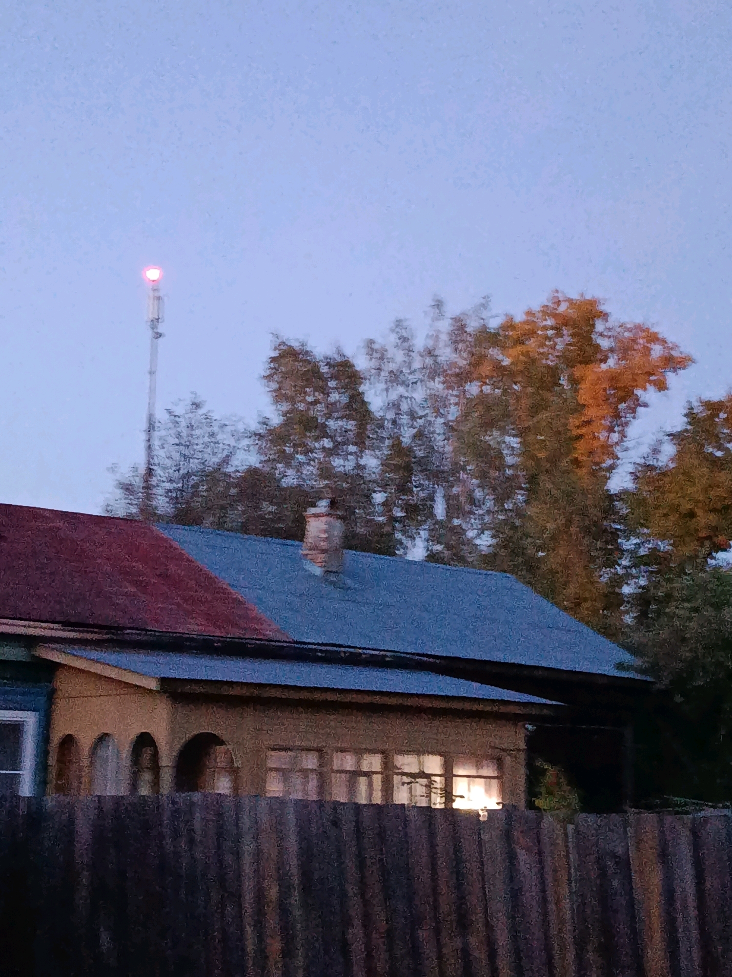 The image captures a twilight scene in the city of Vereya, Russia, featuring a residential building with a prominent red and blue roof. A tall, slender communication tower with a red light at its apex stands in the background, piercing the pale blue sky. Adjacent to the tower, trees display autumn foliage, with some branches illuminated in warm orange hues. The house itself has a porch with arched openings and windows that glow with interior light, suggesting occupancy. A weathered wooden fence runs across the foreground, partially obscuring the lower section of the building. The overall atmosphere is one of a quiet, rural evening.