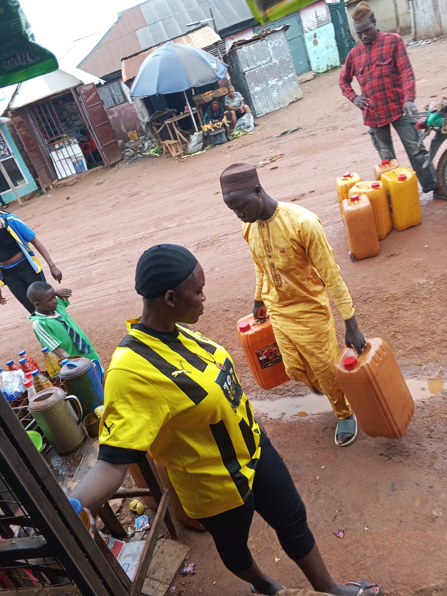 This image depicts an outdoor scene in a marketplace, likely in Abuja, Nigeria. The ground is dirt and appears wet, suggesting recent rain or damp conditions. In the foreground, a woman wearing a yellow and black striped shirt and black leggings is shown from the waist down. Beside her, a young boy in a green shirt with a striped tie is walking. Further back, a man in a yellow traditional outfit is carrying two large orange jerrycans. Another man in a red and black plaid shirt is standing near a motorcycle and several yellow and orange jerrycans are placed on the ground. In the background, there are market stalls with corrugated metal roofs, some selling goods, and a blue umbrella providing shade.
The activity appears to be commerce, with individuals transporting goods, possibly fuel or water, given the presence of the jerrycans. The expressions on the faces of the people are not clearly discernible, but the overall atmosphere seems to be one of daily market activity. The lighting suggests it is daytime, though the weather might be overcast or recently rained. The presence of the marketplace, the attire of the people, and the types of goods visible are consistent with a West African setting. The phrase "Viva" is partially visible on a green awning in the upper left corner.
