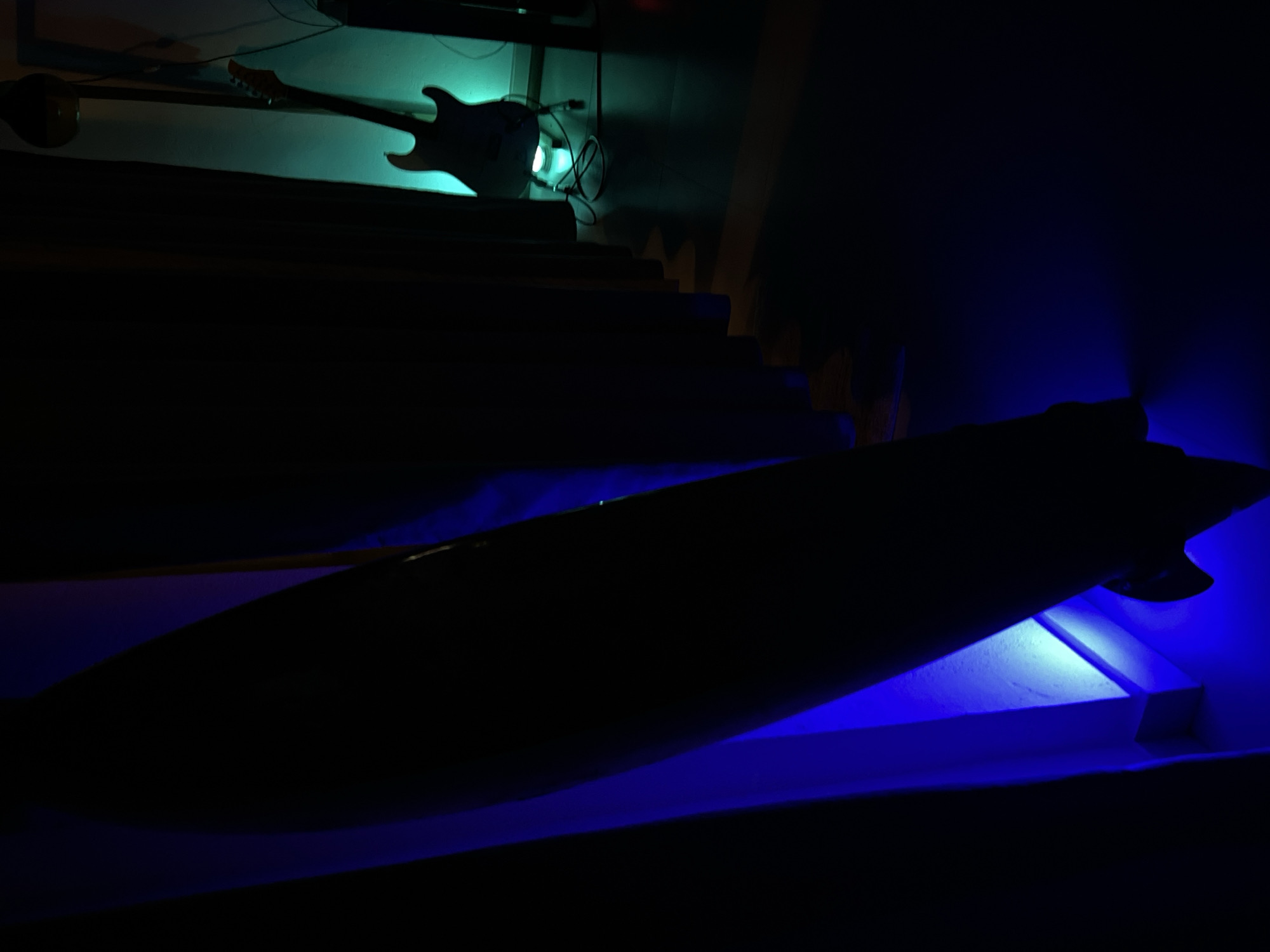 The image presents a dimly lit room, possibly in Goviyapana, Sri Lanka, though this is not definitively confirmed from the visual data alone. A guitar, illuminated by a green light, hangs on a wall above a staircase. Below the staircase, a surfboard is propped up, lit by a blue light. The rest of the room is mostly obscured in shadow.