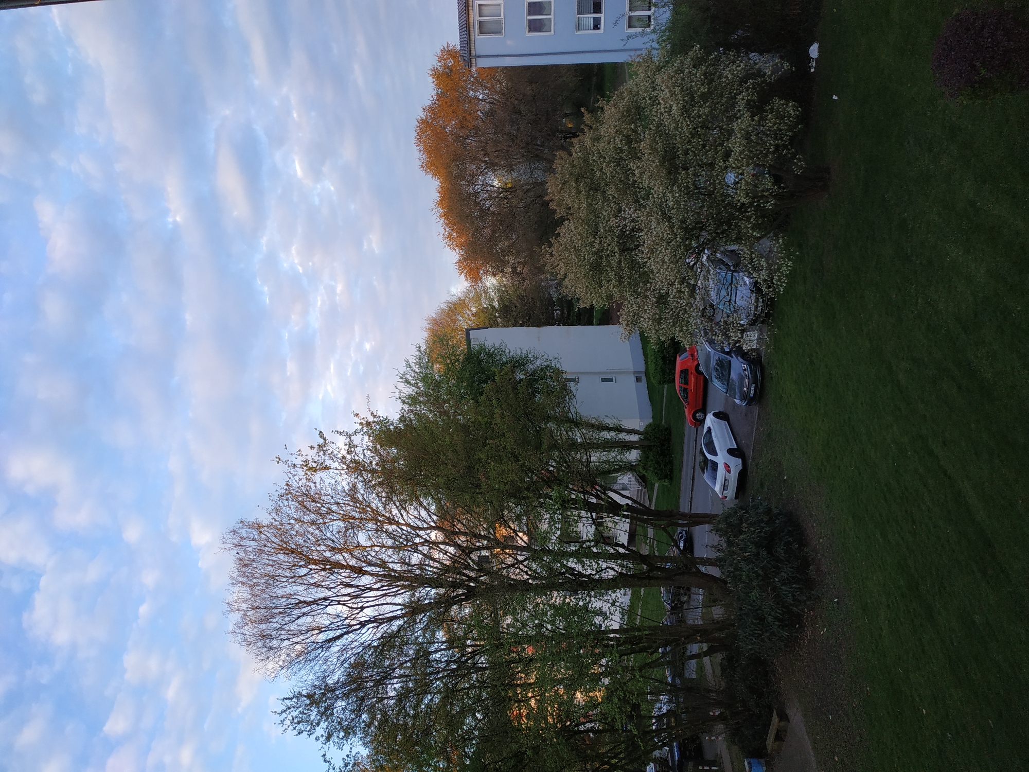 The image shows a view of a residential area in Dortmund, Germany, likely taken from an elevated position, possibly a high-rise apartment. The sky is partially cloudy with blue patches. Several apartment buildings are visible in the background. Trees are prominent, some with green leaves and others with yellow/brownish leaves. Cars are parked along the street adjacent to a grassy area. The environment is typical of a suburban or residential area in Germany.