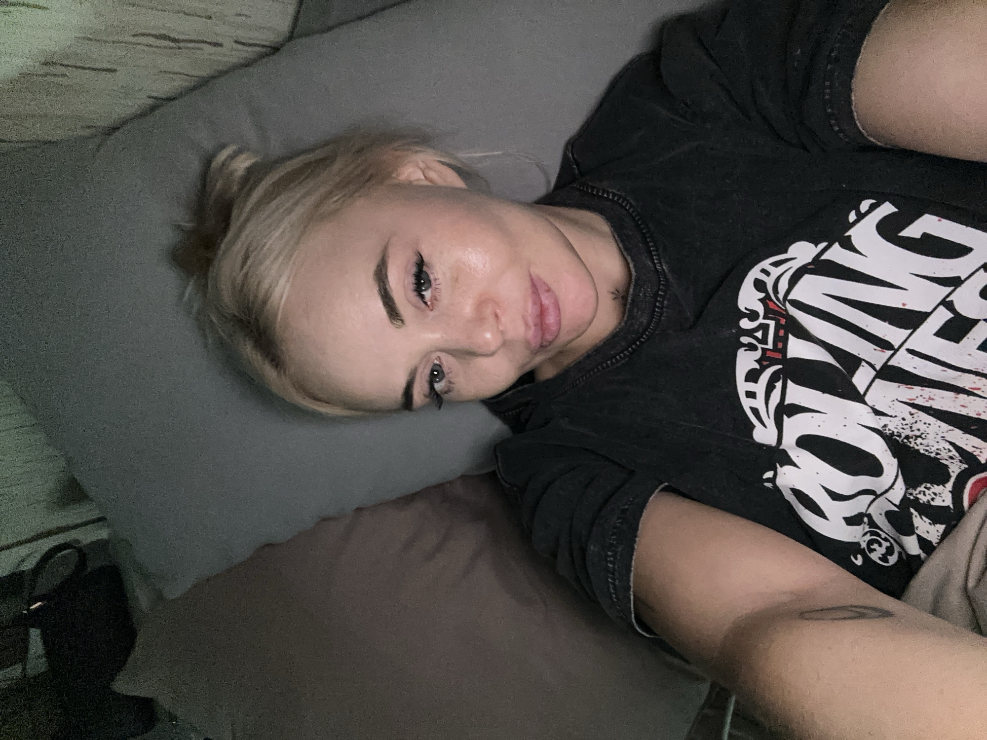 A blonde-haired woman is lying on a bed in a room. She is resting on a gray pillow, with a brown blanket visible below it. She is wearing a black t-shirt with a large white graphic print on the front. Her head is tilted towards the left side of the frame, and she is looking at the camera. A small tattoo is visible on her neck, and another on her left arm. The background shows a wall with a textured surface, possibly wood paneling or wallpaper. A black bag is visible on the left.
The city of Высокий, Russia, is not discernible from this image.
