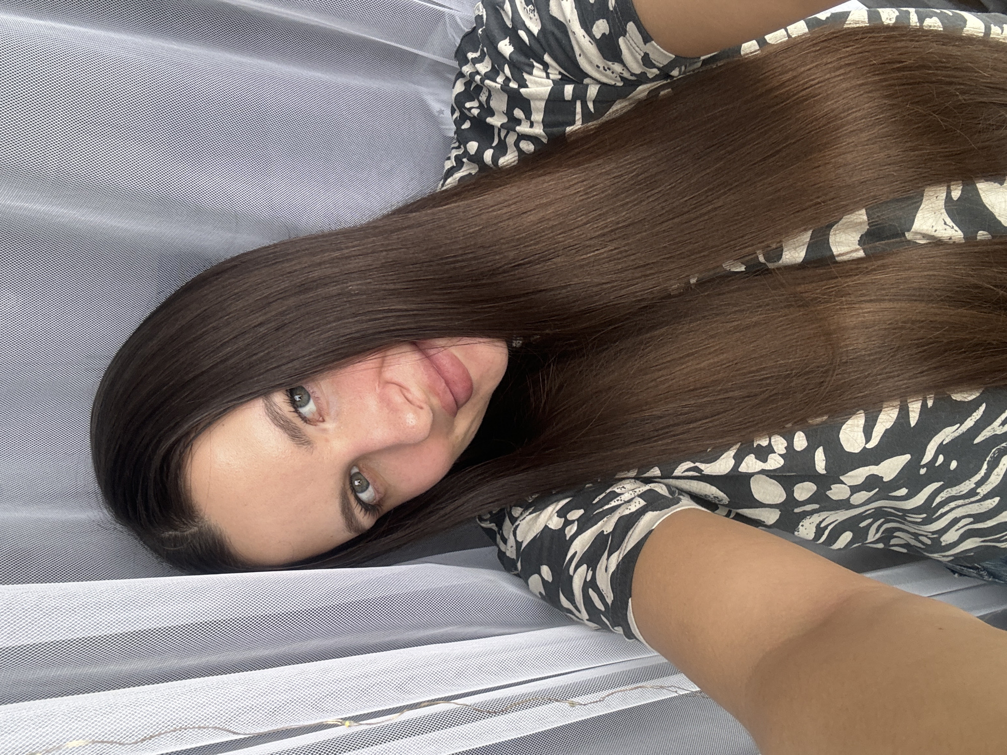An eye-level selfie captures a young woman lying down in Akulovo (Gp Kubinka), Russia, looking directly at the camera with a subtle smile and light-colored eyes. Her long, straight, dark brown hair cascades across a significant portion of the frame, partially covering her torso. She is dressed in a top or dress featuring a distinctive black and white abstract or animal-print pattern.
The woman's arm is visible in the lower right, suggesting she is holding the camera for the selfie. The background primarily consists of a light, sheer, mesh-like fabric, possibly a net or gauze, which is draped with gentle folds and exhibits a fine grid pattern of small holes. Faint, warm-toned string lights are visible peeking through this translucent fabric in the lower-center of the image, casting a soft glow. The overall lighting is gentle and diffuse, likely indicating an indoor setting. There is no visible text in the image.