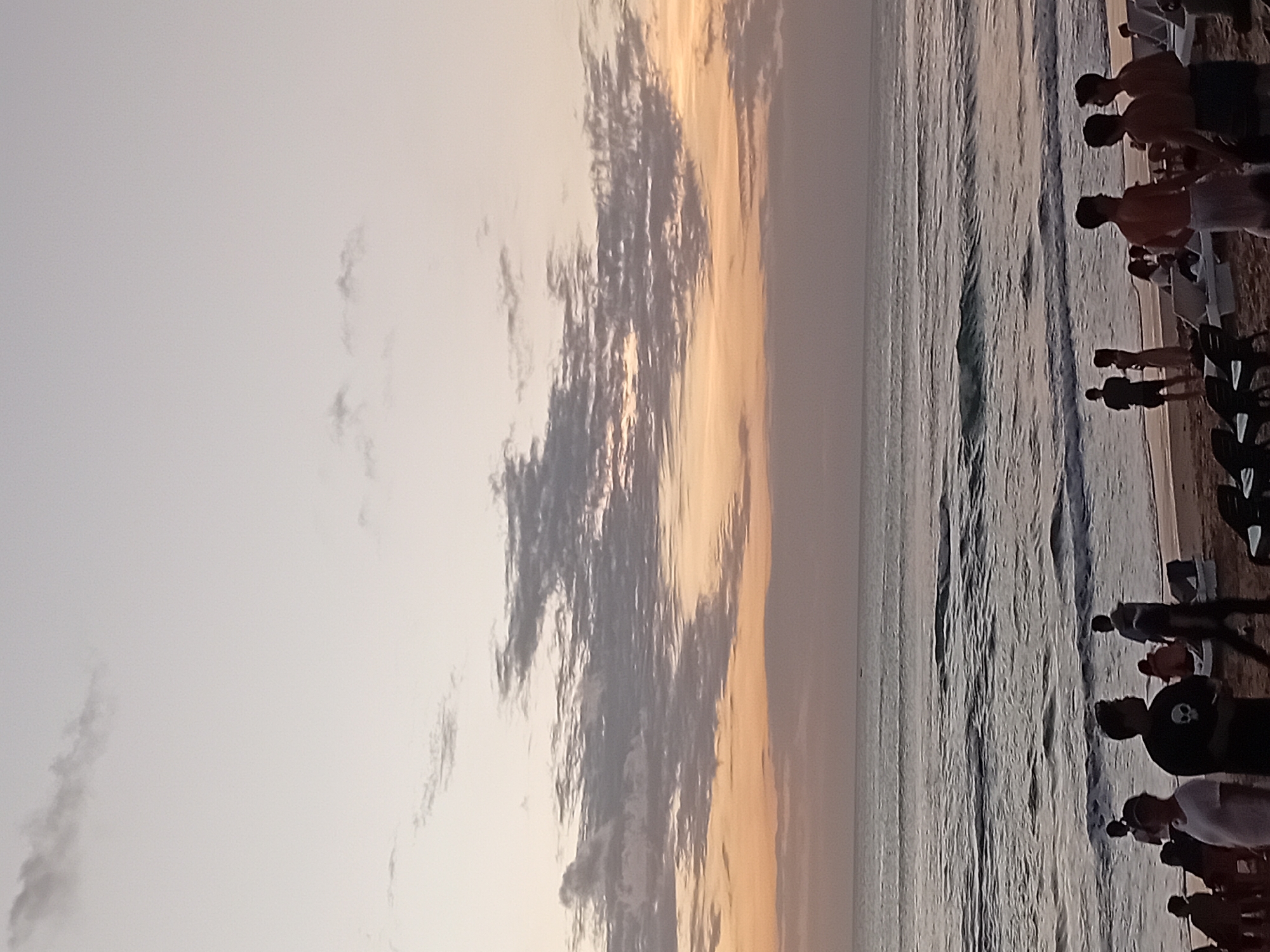 This image depicts a serene beach scene in Kuta, Indonesia, during a dusk or dawn. The sky is painted with soft hues of orange, peach, and grey, with scattered clouds creating a textured panorama above the calm ocean. Gentle waves roll onto the sandy shore where a group of people are gathered. Some individuals are walking along the beach, while others are seated on beach chairs or engaging in conversation. The atmosphere is tranquil, suggesting a peaceful evening by the sea.