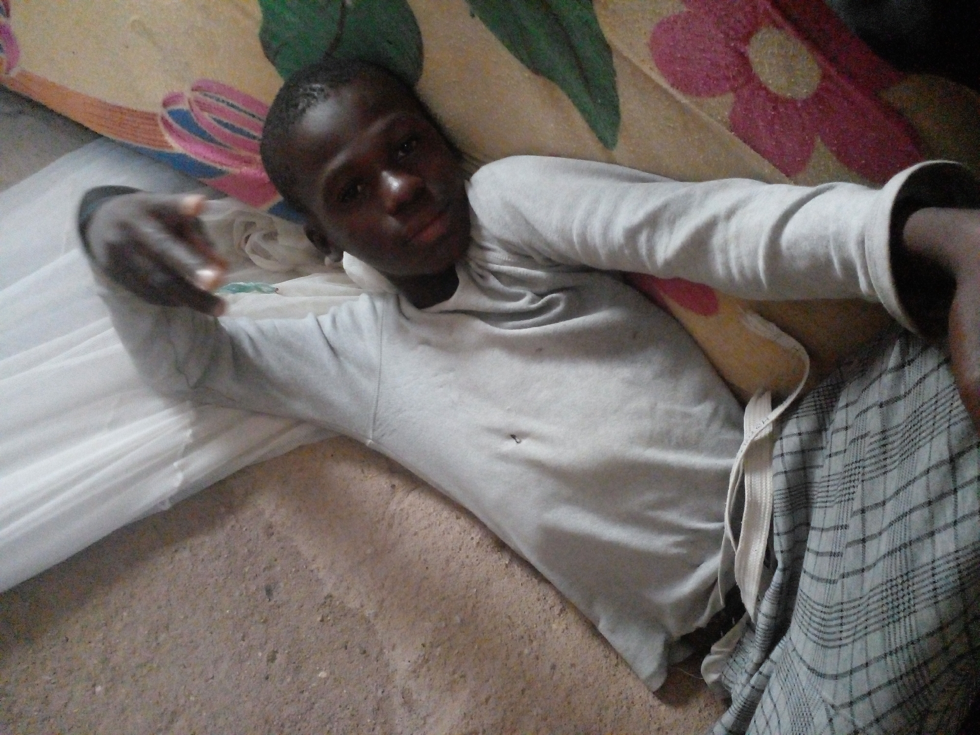 A young person with dark skin is lying on their back on a patterned blanket. They are wearing a light gray long-sleeved shirt and plaid pants. The person's right arm is raised and bent at the elbow, with their hand positioned in front of their face. Their head is resting on the blanket, and they are looking towards the camera. The background consists of a concrete floor and a white fabric. The scene appears to be indoors.