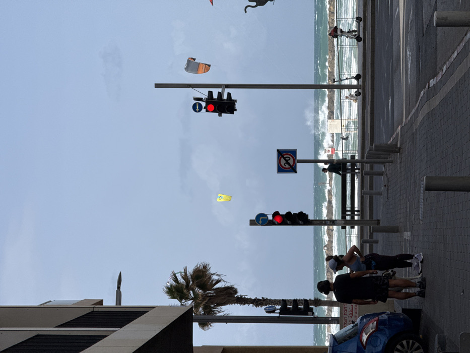 The image depicts a vibrant coastal urban scene in Tel-Aviv, Israel, overlooking the Mediterranean Sea. The sky is partially overcast with patches of lighter blue. The sea is turbulent, characterized by large, white-capped waves crashing against a rock barrier that extends along the coastline.
In the immediate foreground, a paved sidewalk occupies the bottom portion of the frame. On the left, a blue hatchback car is parked. Adjacent to it, a male individual in a dark t-shirt, dark shorts, and headphones stands with a female in a light tank top and dark leggings. A light-colored, medium-sized dog is positioned between them. Both individuals appear to be facing towards the right, engaging in a paused interaction.
To their right, a paved road runs parallel to the sidewalk, marked by several dark grey bollards. Beyond the road, a wide promenade stretches along the beachfront. Two dark traffic light poles with illuminated red signals are prominently positioned on the promenade, accompanied by a blue circular 'turn left' arrow sign and a square blue sign indicating a '20' speed limit or restriction.
On the promenade, two dark benches face the sea. A lone individual in dark clothing stands near one bench, observing the water. Further right, another person is seen riding an electric scooter towards the right of the frame, while a second electric scooter is parked unattended. A person is discernible within the breaking waves near the rock barrier.
In the mid-ground, a tall palm tree stands on the left, next to a multi-story building with visible balconies. High above the sea, two colorful kite surfing kites are airborne: one yellow on the far left and another orange and grey on the right, indicating active water sports and windy conditions. A dark, stylized sculpture resembling a bird or mythical creature is partially visible on the far right, near the edge of the promenade.