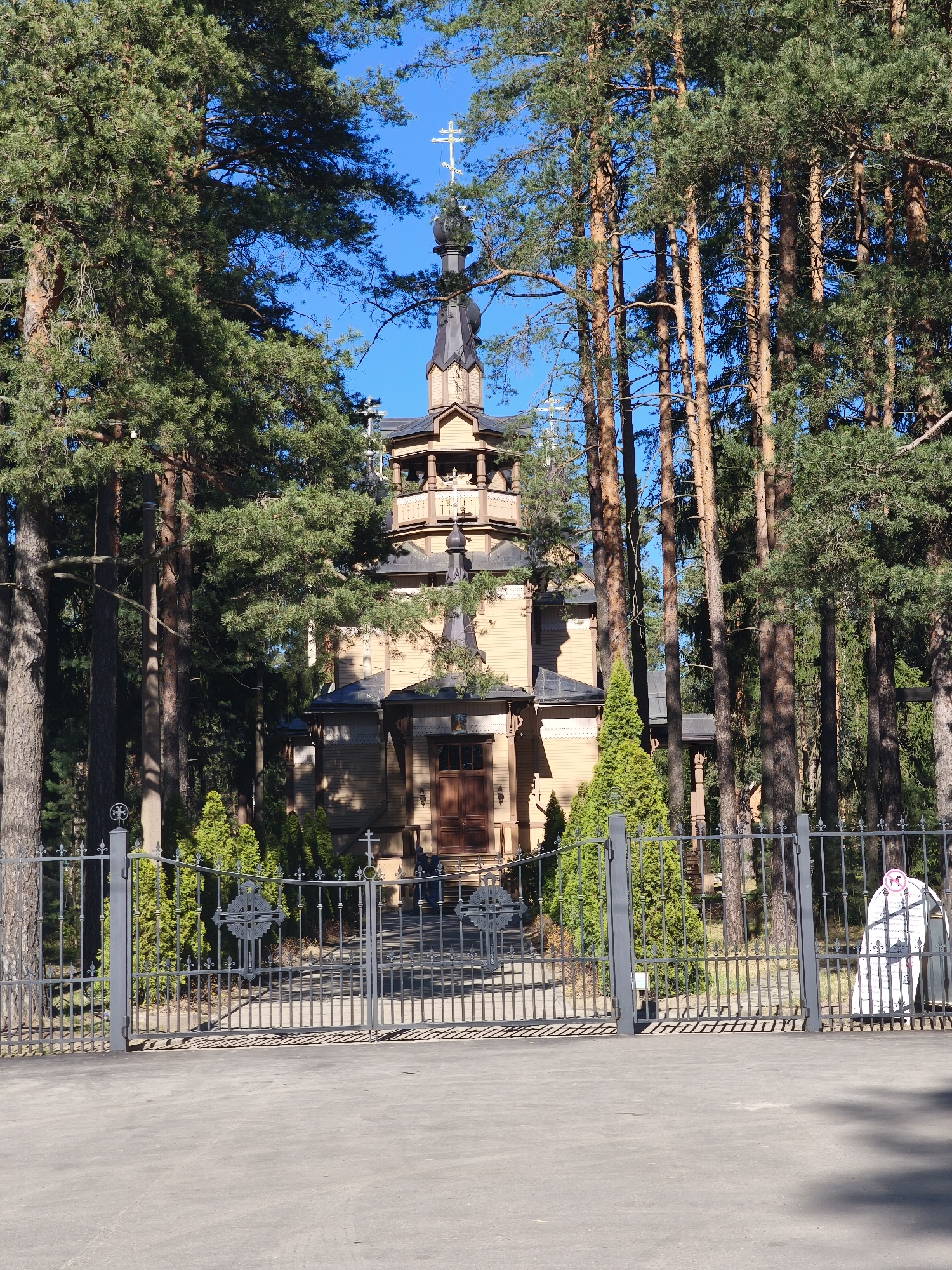 The image shows the wooden Church of St. Sergius of Radonezh in Pesochny, Russia. The church is surrounded by tall pine trees. A gray metal fence with ornate Christian crosses forms the entrance. The pavement in front of the gate leads to the church. A sign is visible to the right of the gate. The church itself is multi-tiered with a dark spire topped with a cross. The sky is clear blue.