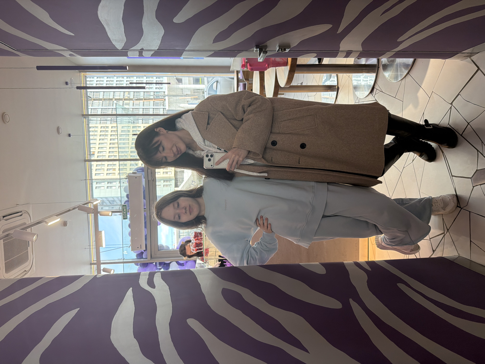 Two young women pose in a reflective setting with purple zebra-striped walls. The upper woman holds a smartphone, wearing a brown coat over a light-colored hoodie, black leggings, and black Mary Jane shoes. The lower woman wears a light blue sweatsuit and white sneakers, posing with one hand on her hip. Behind them are windows revealing buildings, possibly in Perm, Russia, based on prior knowledge. The interior includes tables and chairs, and there are purple decorations near a window. The reflection creates a mirrored effect, presenting the scene from multiple perspectives.