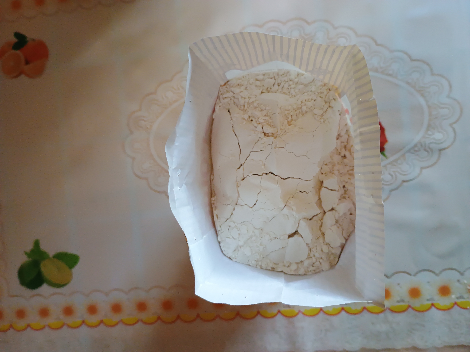 The image shows a bag of flour on a tablecloth. The tablecloth has a pattern of flowers and fruit. The flour is light brown and has a cracked surface. The bag is open at the top, and the flour is spilling out. The image is taken from a high angle, looking down at the bag of flour. The lighting is natural, and the image is in focus. The location in the city of Zaporizhzhia, Ukraine, is not specified.