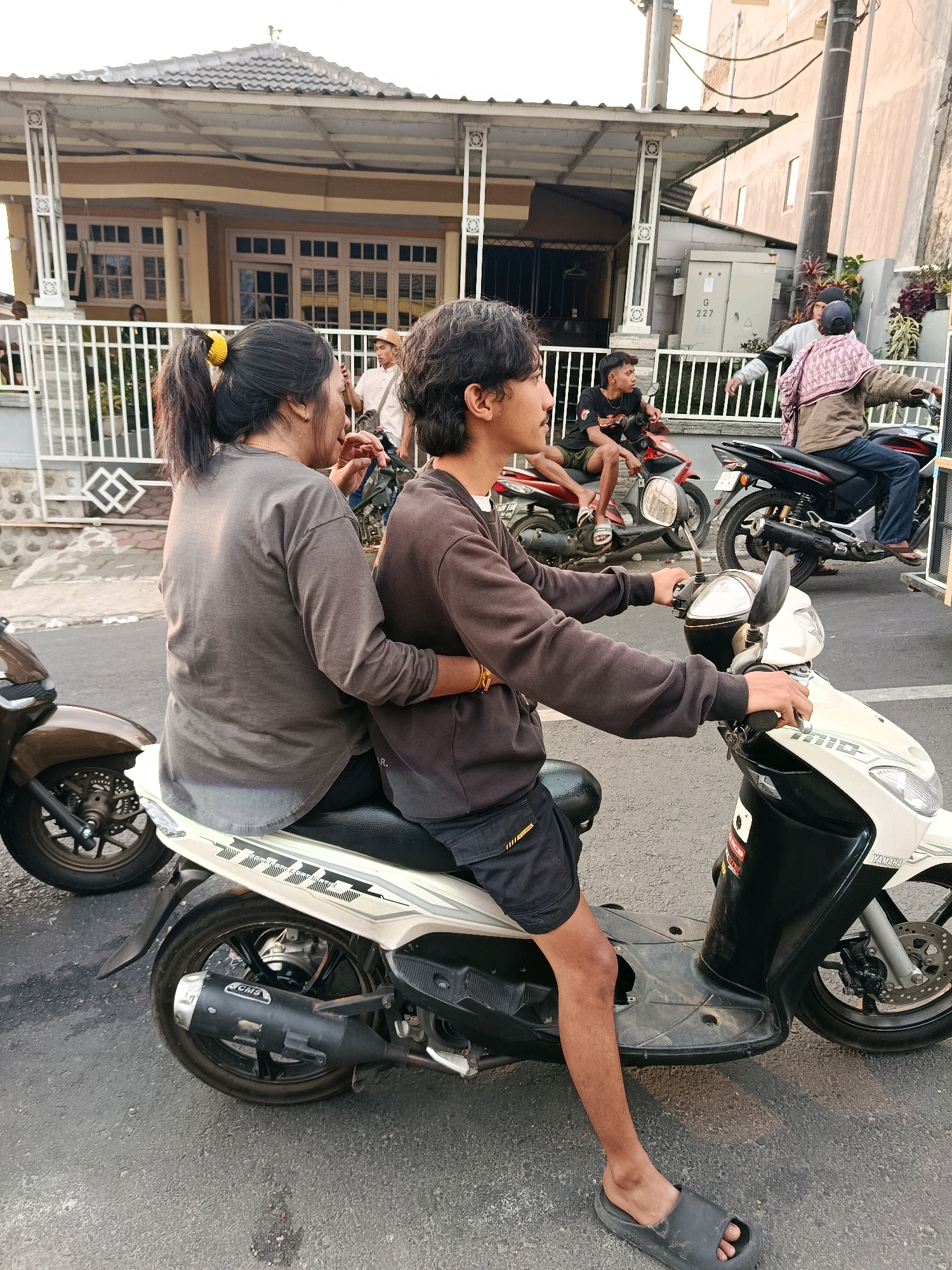 This is a medium shot, eye-level, outdoor photograph of two individuals riding a white and black scooter on a paved road in Gubukklakah, Indonesia. The scooter is in the foreground, angled to the right. The rider, a young male with dark, wavy hair, wears a brown long-sleeved shirt and black shorts. His hands are on the handlebars, and his left leg is visible, wearing a black slide sandal. The passenger, a female with dark hair tied in a ponytail with a yellow elastic band, is seated behind the rider and wears a grey long-sleeved shirt and dark pants. Her right arm is around the rider's waist.
In the background, several other people and scooters are visible. A red scooter with two young males is parked or moving to the left. Further back, a black scooter with a male rider wearing a blue cap and a patterned cloth draped over his shoulders is also present. A building with a light-colored facade and a white railing is situated behind the people and scooters. The environment suggests a street or roadside setting, with overhead wires and a utility box visible. The lighting indicates it is daytime, with the sun casting soft shadows.