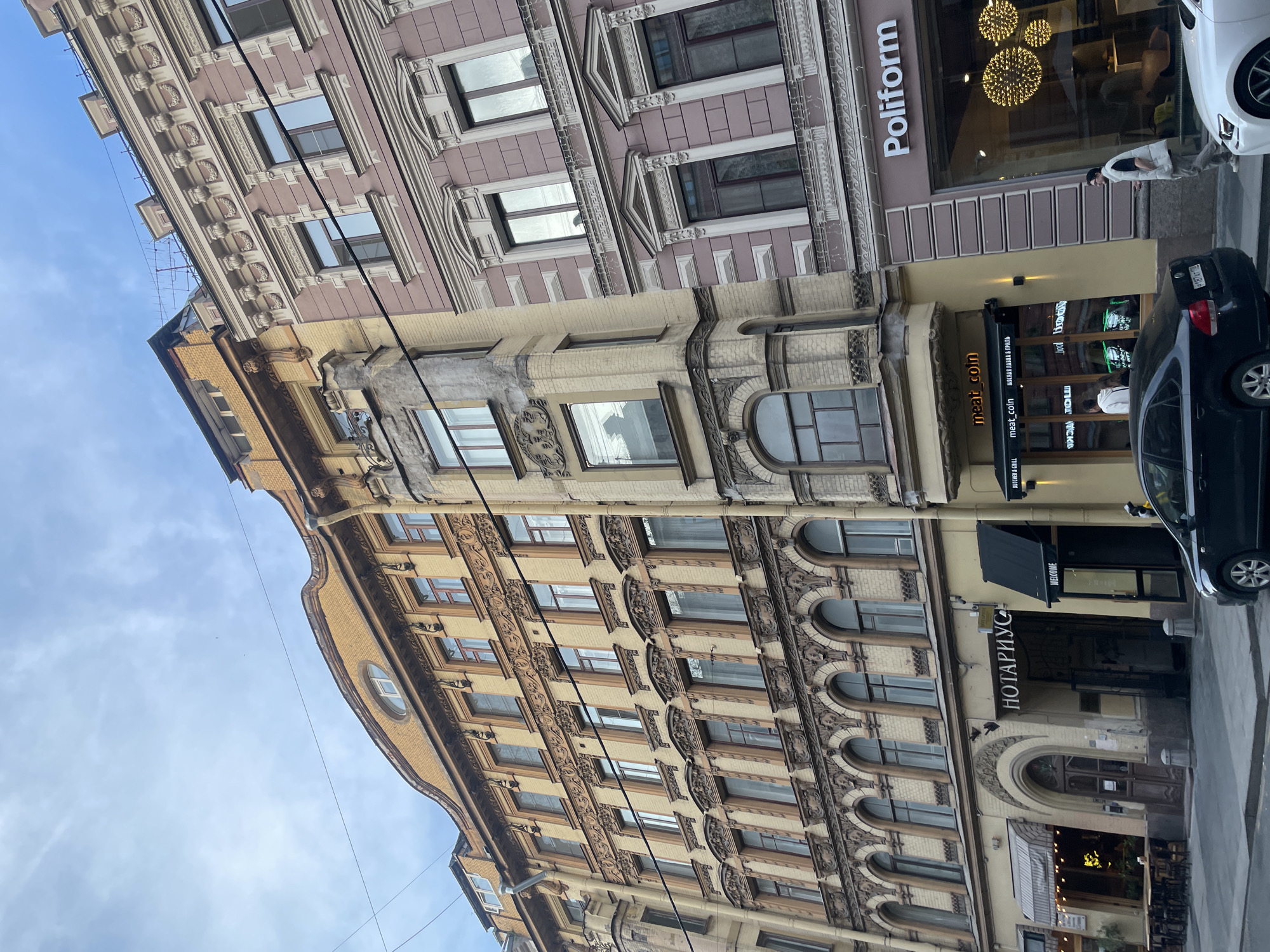 An outdoor, eye-level shot captures a vibrant street scene in Saint Petersburg, Russia, showcasing two distinct multi-story buildings with ornate facades under an overcast, bright blue sky. The building in the lower and middle parts of the frame is a grand, beige-yellow structure with intricate architectural details, including decorative window frames, arched windows, and sculptural elements. Adjacent and above it, another building with a pinkish-mauve facade features rectangular windows, cornices, and decorative stucco work. A thin black power line stretches across the scene.
At street level, several businesses operate from the ground floors of these buildings. The pinkish-mauve building hosts a "Poliform" store, likely a furniture or design showroom, with large display windows revealing spherical light fixtures inside. Below, on the beige-yellow building, a restaurant named "meat_coin" is visible, with additional signage reading "BUTCHER & GRILL" and "МЯСНАЯ ЛАВКА В ГРИЛЬ" (Meat Shop in Grill). Further down the street, an office is identified by the Russian sign "НОТАРИУС" (Notary) with "WELCOME" beneath it.
Two cars are parked along the curb: a black hatchback and the front portion of a white car. A person in a light-colored vest and pants is walking on the sidewalk near the white vehicle. The scene depicts a lively urban environment, characteristic of a European city, under natural daylight conditions.