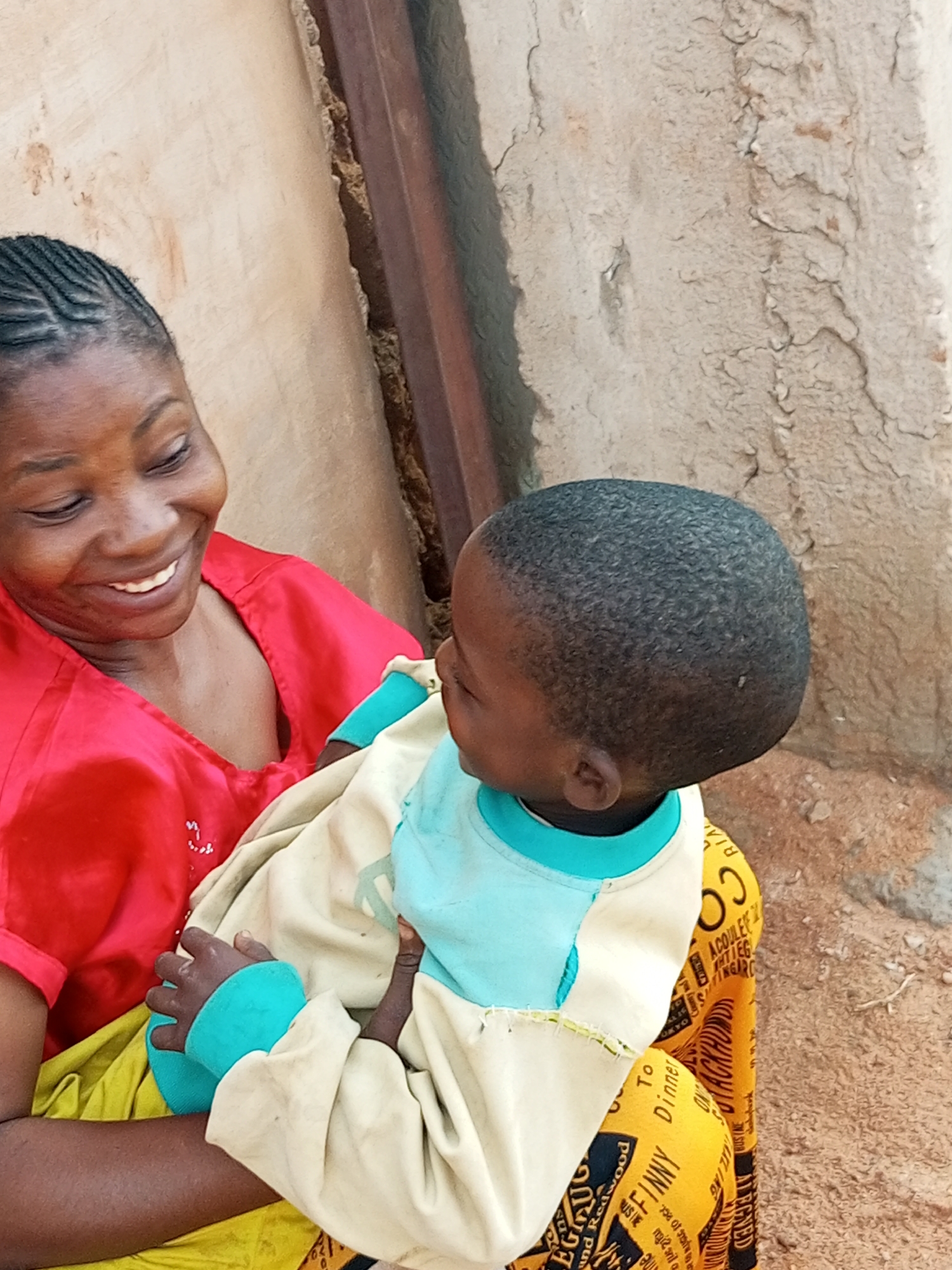 A woman with braided hair smiles warmly as she holds a young child swaddled in blankets and wearing a blue-collared shirt. The child is turned away from the camera, with their head tilted down. The woman is wearing a vibrant red top and a yellow skirt. They are seated outdoors, with a textured, light-colored wall behind them and a wooden structure to the right. The ground appears to be dirt. The context of a city in Unknown, Unknown is provided.