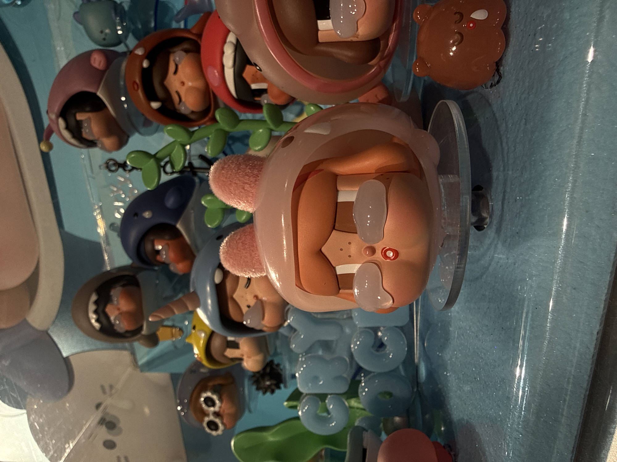 This image depicts a close-up view of a display of collectible figurines, likely from a toy store or exhibition. The figurines are small, stylized characters with exaggerated features, many wearing themed outfits such as shark costumes and bunny ears. Some have tear-like embellishments on their faces, suggesting a "crybaby" or sad theme. The overall color palette is bright and cheerful, dominated by pastel blues, pinks, and oranges, with a sparkling, translucent blue surface acting as the display base. In the background, other larger, softer-looking figures and decorative elements are visible. The words "CRY OCGI" are spelled out in large, translucent blue block letters in the mid-ground. A small, brown, bear-like figurine with closed eyes and a single tear is positioned in the lower right corner. The scene appears to be an indoor retail or entertainment environment. There is no indication of the city of Pudong, China.