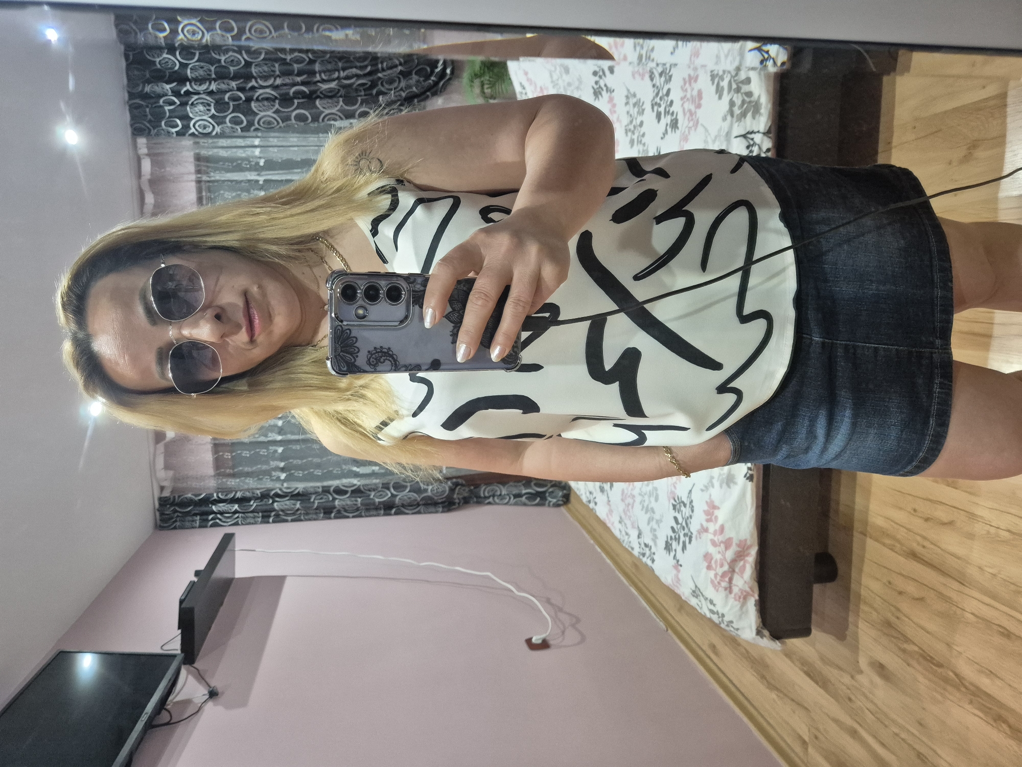 A woman is taking a mirror selfie in a bedroom. She has blonde hair and is wearing sunglasses, a white and black patterned tank top, and a denim skirt. Her phone is visible in her hand, as is a tattoo on her shoulder. The bedroom has a bed with a floral comforter, a television mounted on the wall, and curtains. The floor is made of wood.