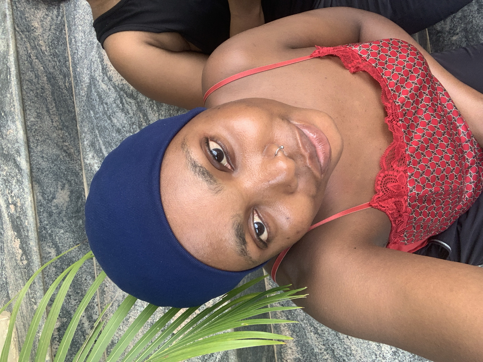 An overhead, close-up shot captures a young Black woman wearing a dark blue beanie and a red and black patterned camisole. She is looking directly at the camera with a subtle smile. Her skin is dark, and she has a nose ring. To her left, a vibrant green palm frond extends into the frame. In the background, a person wearing a black tank top is visible, and the ground appears to be made of large, grey, textured stones, possibly steps. The scene is outdoors.