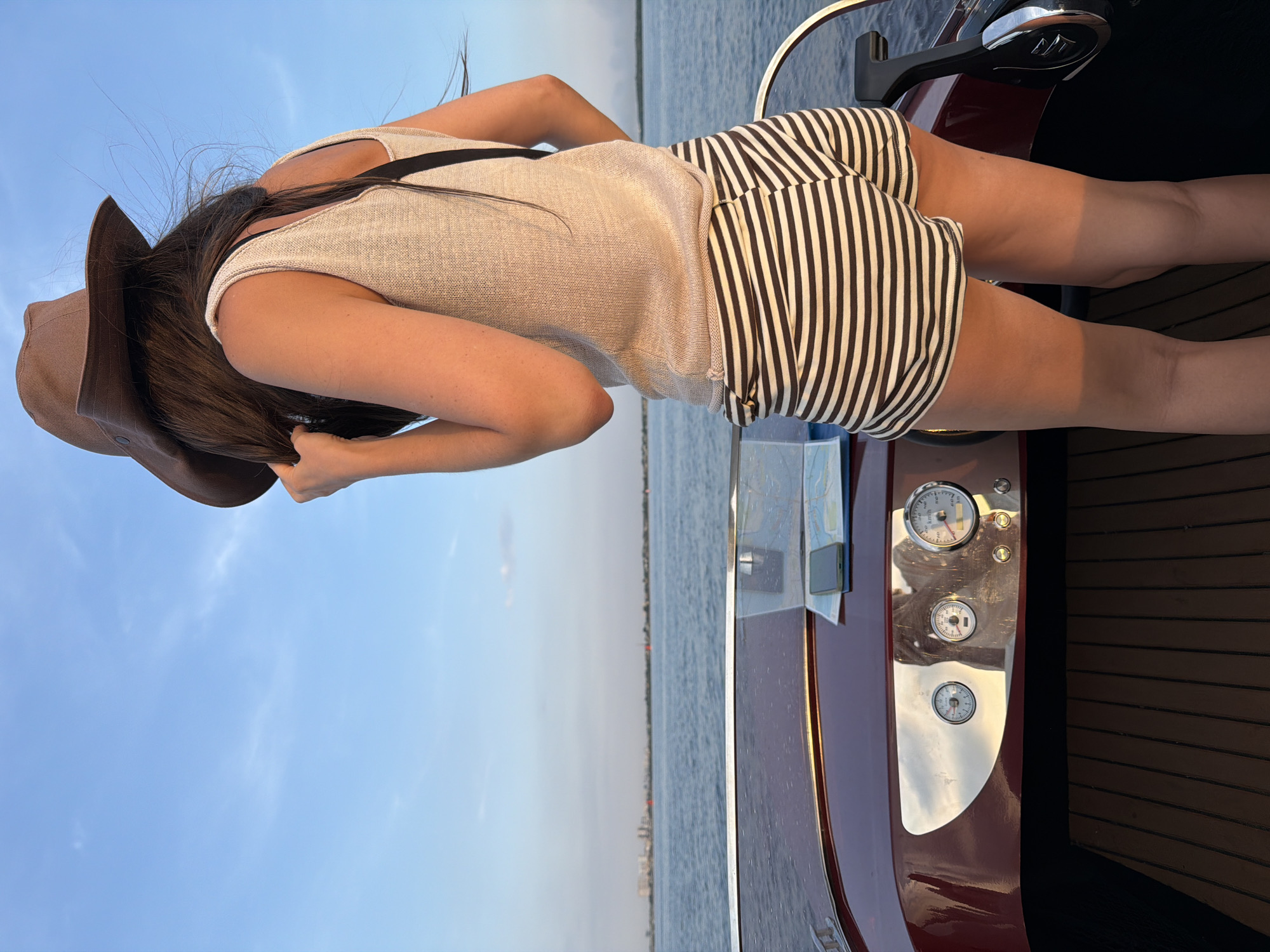 A woman wearing a brown hat, a beige knit tank top, and striped shorts stands on the deck of a boat. She is holding her hair back with her right hand. The boat has a dark red dashboard with gauges and a steering wheel. A map and a smartphone are on the dashboard. The boat is on a body of water, likely a river, with a city skyline in the distance. The sky is blue with a few wispy clouds. The lighting suggests it is either sunrise or sunset. The location is the city of Saratov, Russia.