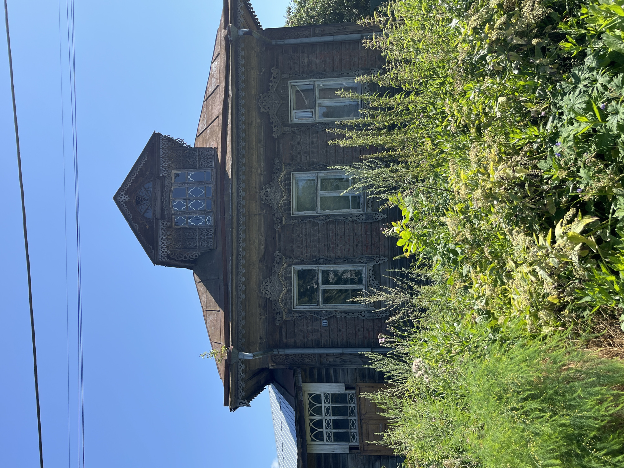 The image depicts an old wooden house with ornate carvings and shutters, located in Kladovitsy, Russia. The house has a steep, rusty metal roof and a dormer window with decorative woodwork. The facade features several windows, some with intricate patterns. A small balcony with a white-framed window is visible on the left side. The house is surrounded by lush green vegetation and tall weeds, with some wildflowers interspersed. The sky is clear and blue, and power lines are visible running across the top of the image. The house appears weathered, suggesting it is historical. The overall impression is of a rural or semi-rural setting in Russia.